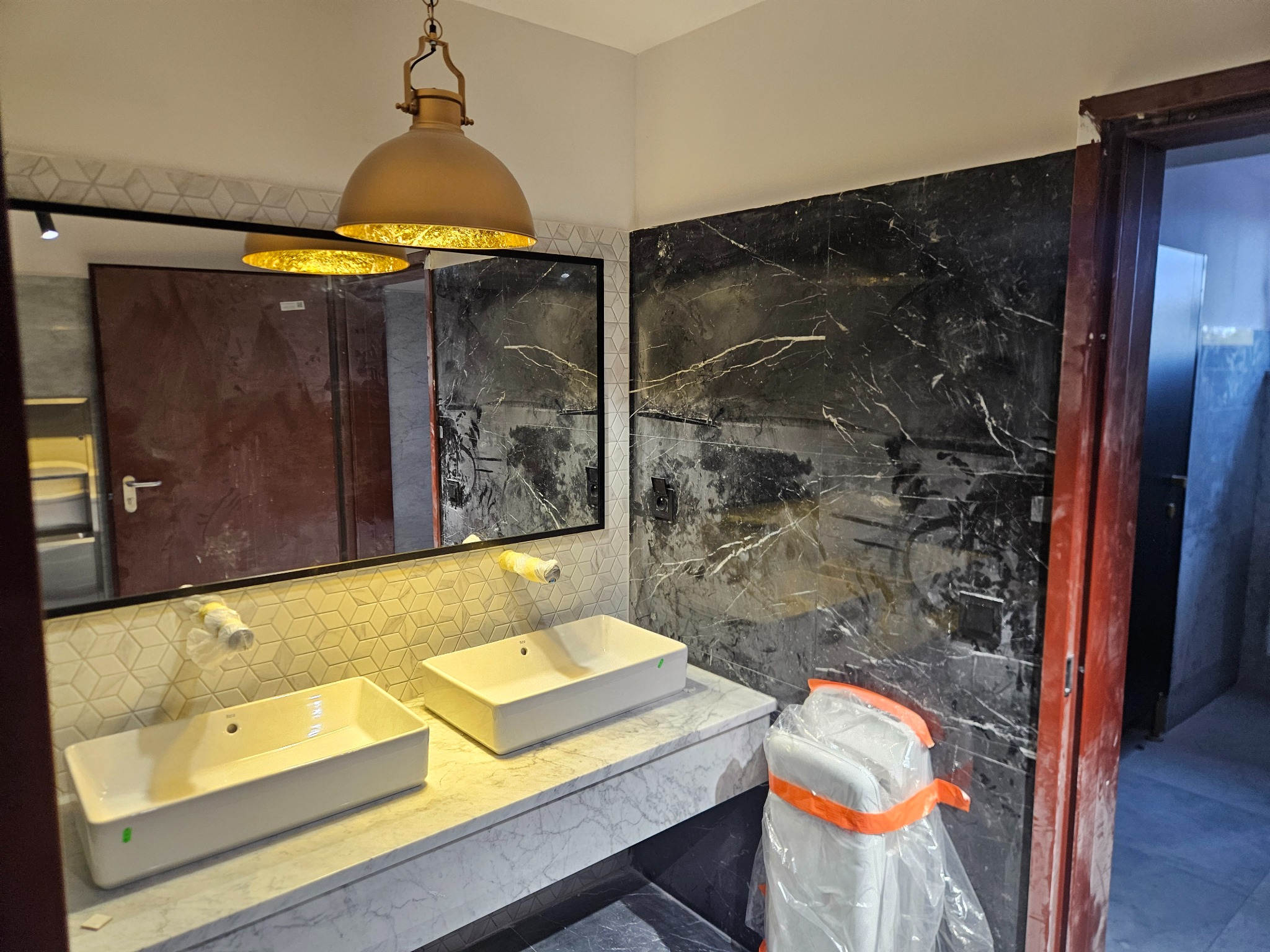 Nowoczesna łazienka z marmurową ścianą i blatem, dwie białe umywalki, złota lampa wisząca. Widoczne zabezpieczenia na armaturze. Drzwi w kolorze terakoty.