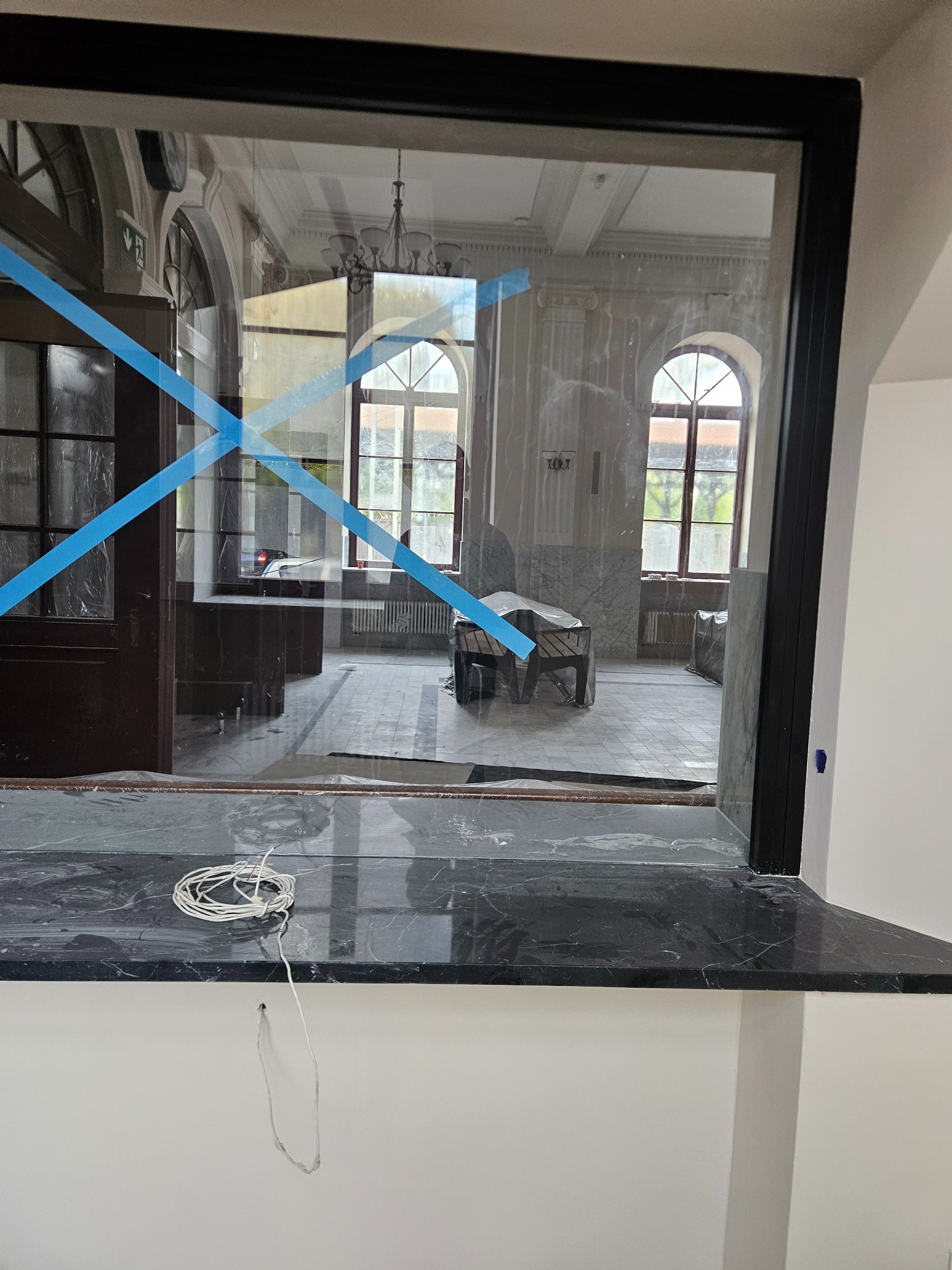 Okienko z czarną ramą i marmurowym parapetem, zabezpieczone taśmą. Widok na wnętrze w trakcie prac, z widocznymi oknami i elementami dekoracyjnymi.