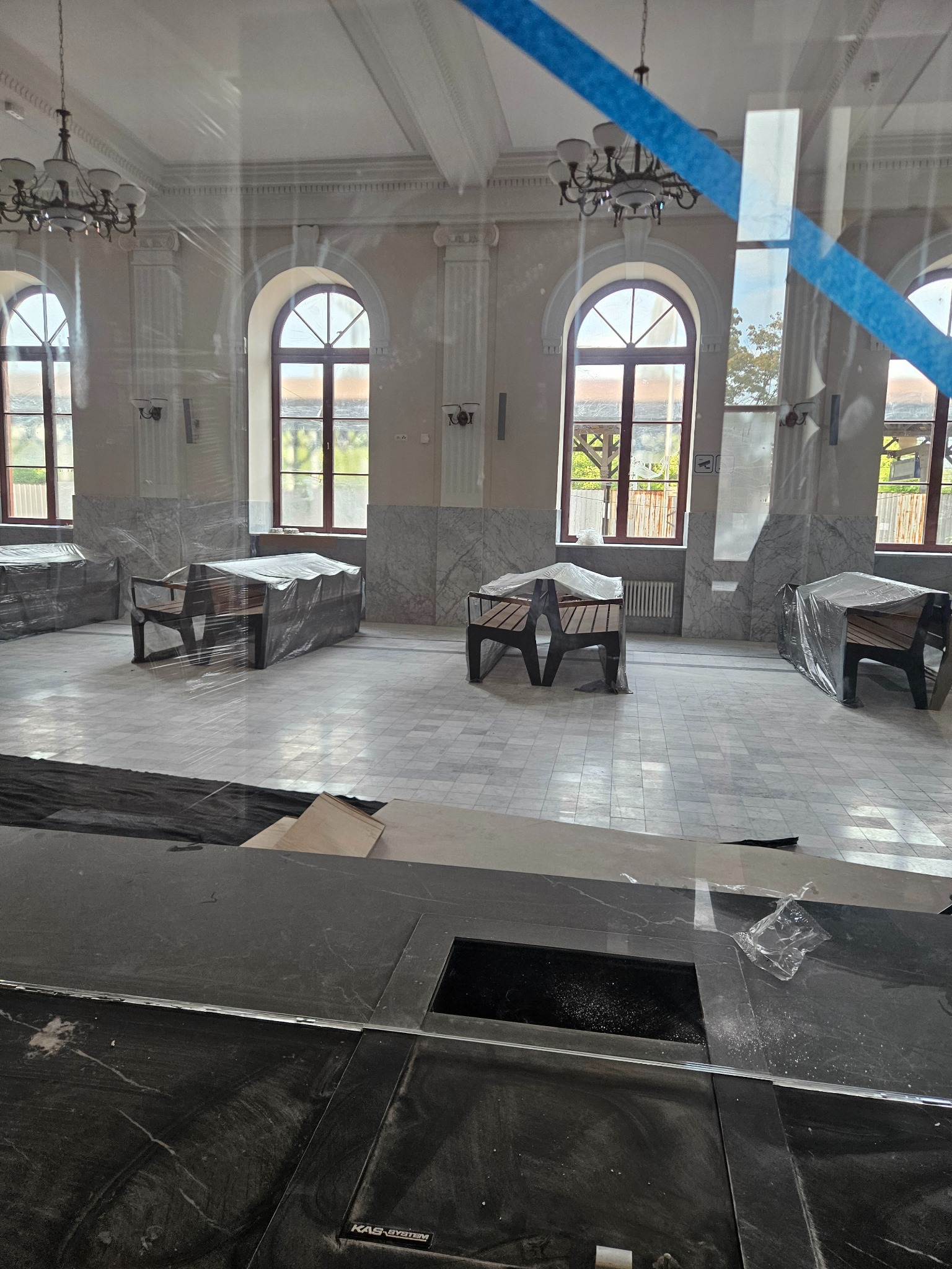 Sala w trakcie remontu: zabezpieczone meble folią, widoczne okna z drewnianą stolarką, marmurowe ściany do połowy wysokości, posadzka z płytek. Przygotowania do malowania.