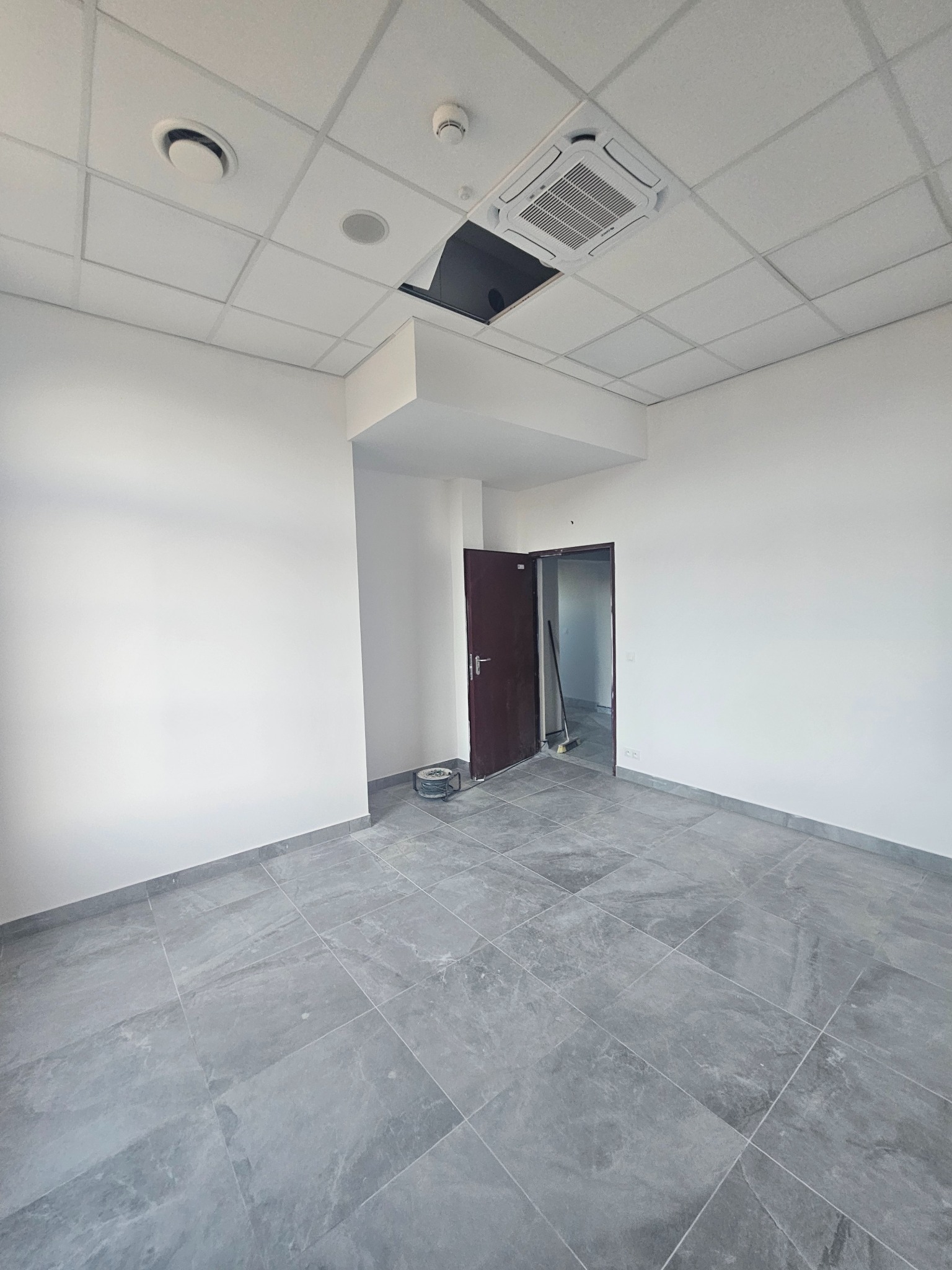 Wnętrze biurowe po remoncie: szare płytki podłogowe, białe ściany i sufit z klimatyzacją. Widoczne narzędzia malarskie. Minimalistyczny design.