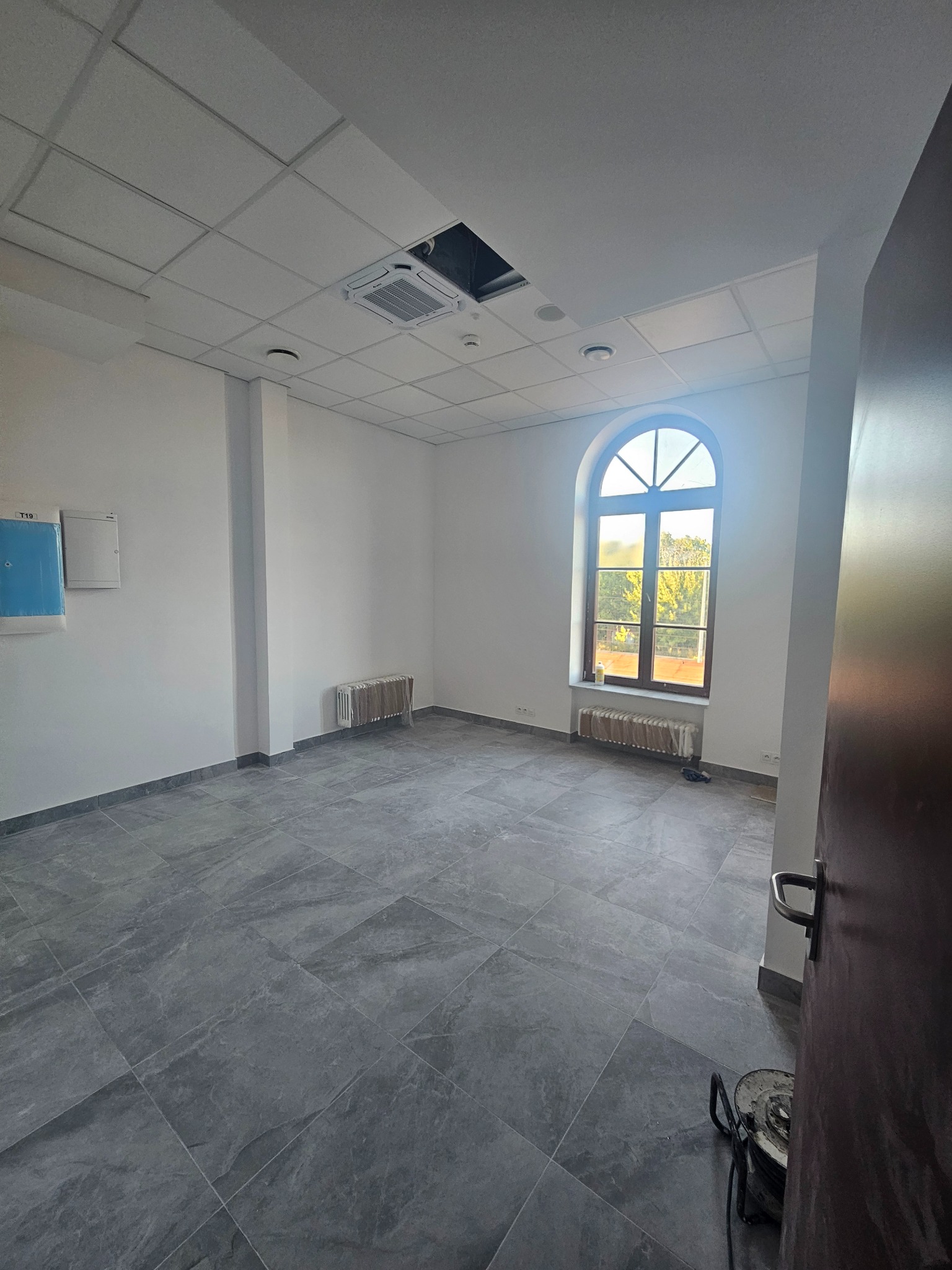 Puste, jasne pomieszczenie biurowe z szarą podłogą, białymi ścianami, oknem łukowym i widoczną instalacją sufitową. Przygotowane do dalszych prac wykończeniowych.
