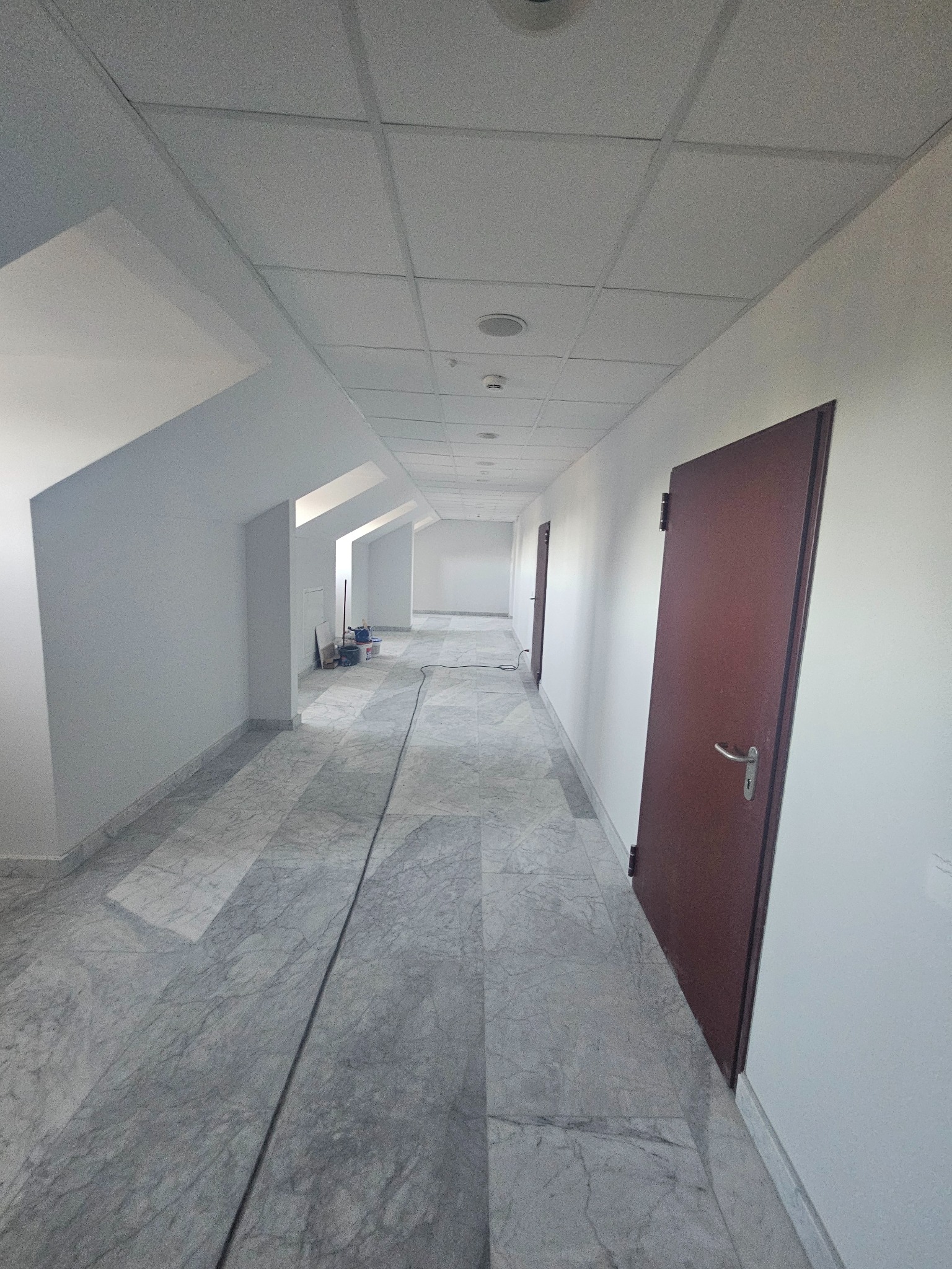 Długi korytarz biurowy po remoncie, z białymi ścianami, szarą marmurową podłogą i brązowymi drzwiami. Widoczne narzędzia malarskie w oddali.
