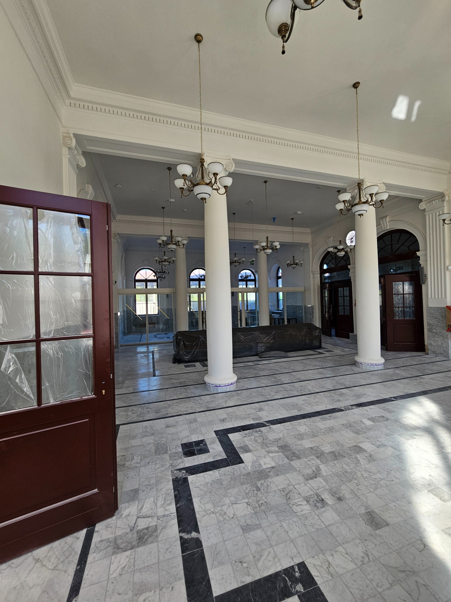 Przestronne wnętrze z marmurową posadzką i kolumnami, widoczne otwarte, drewniane drzwi z szybami zabezpieczonymi folią. Klasyczne lampy wiszące.