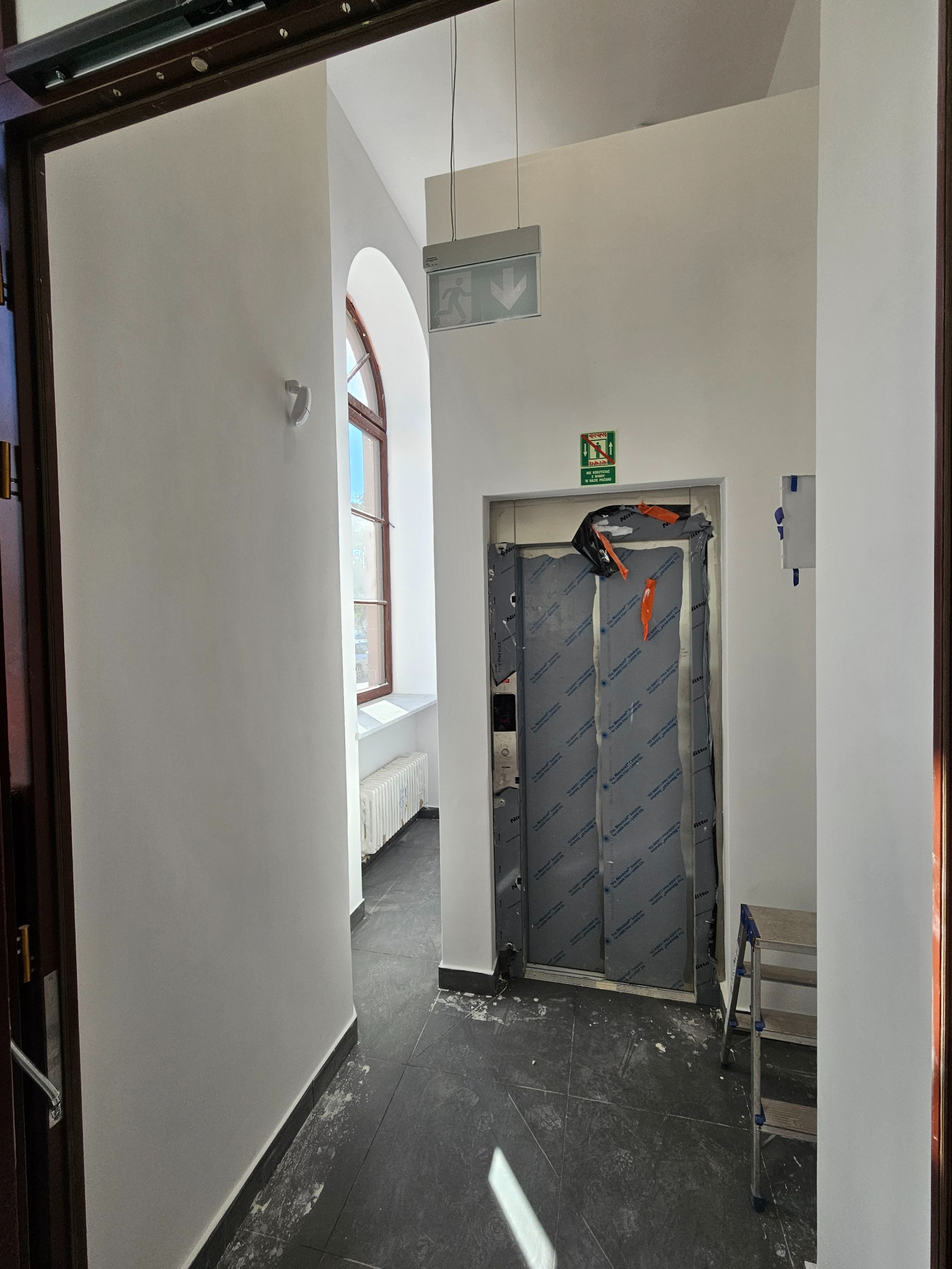 Korytarz w trakcie odświeżania: świeżo pomalowane ściany na biało, zabezpieczone drzwi windy, widoczne narzędzia i drabina. Okno z łukiem doświetla przestrzeń.