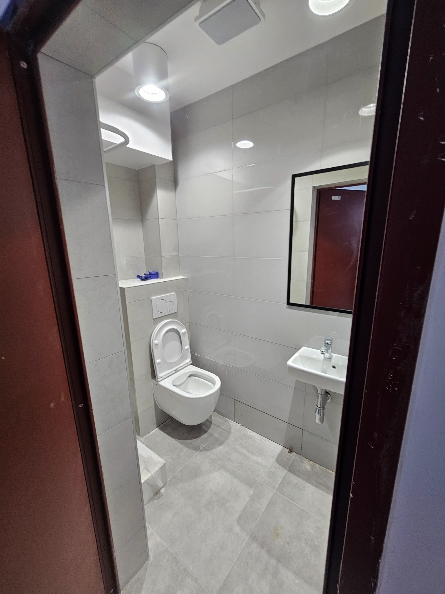 Jasna łazienka z toaletą, umywalką i lustrem w czarnej ramie. Widok przez otwarte drzwi z ciemną ościeżnicą. Minimalistyczny design, szare płytki.