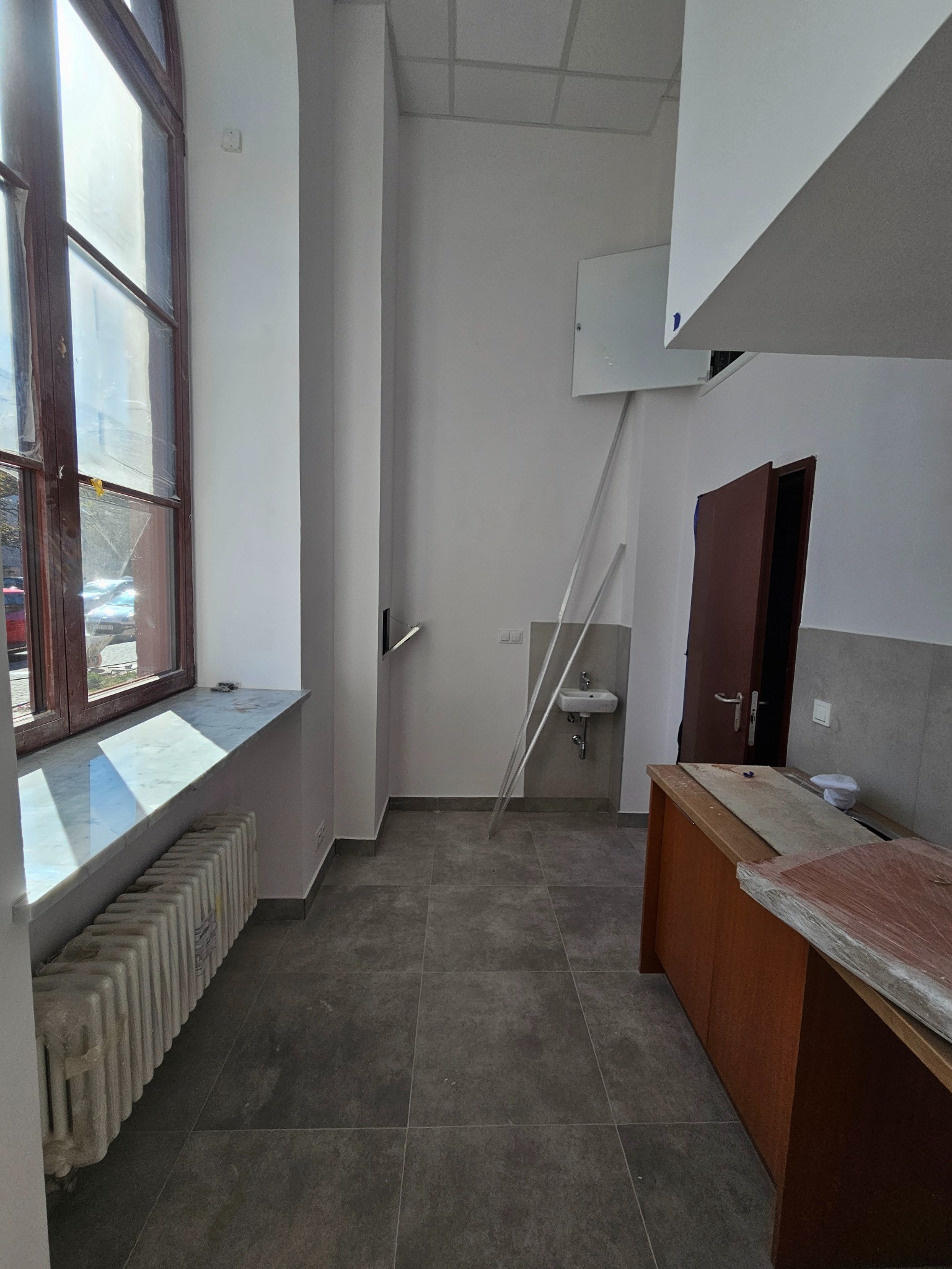 Wnętrze biura przed remontem: grzejnik, okno z marmurowym parapetem, szare płytki podłogowe, umywalka i drzwi. Widoczne ślady użytkowania i przygotowania do prac.