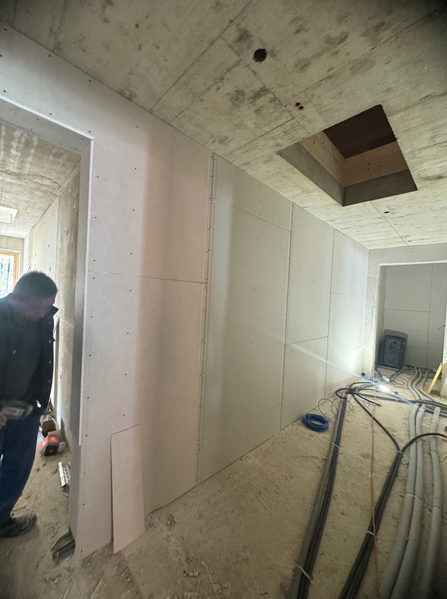 Montaż płyt gipsowo-kartonowych na ścianie w surowym wnętrzu. Widoczne kable elektryczne i otwór w betonowym suficie. Fachowiec w trakcie pracy.