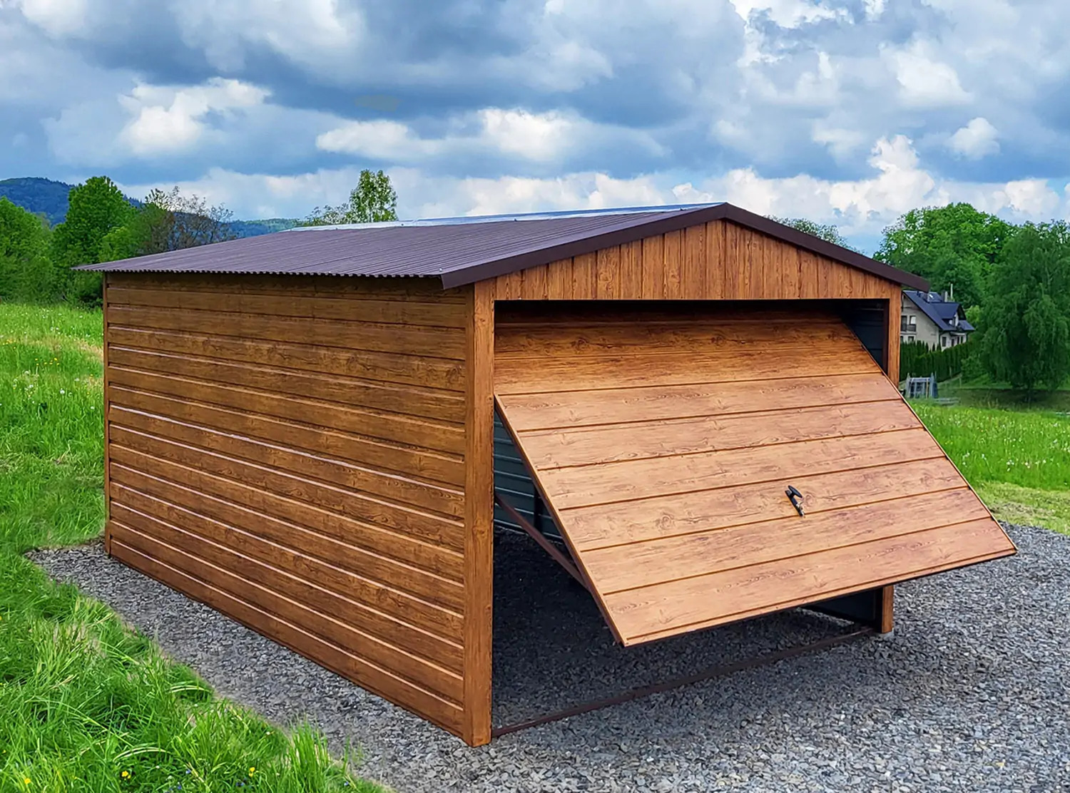 Drewnopodobny garaż z uchylną bramą na żwirowym podłożu, z zielonym tłem wzgórz i błękitnym niebem. Widoczna faktura drewna i metalowe elementy konstrukcyjne.