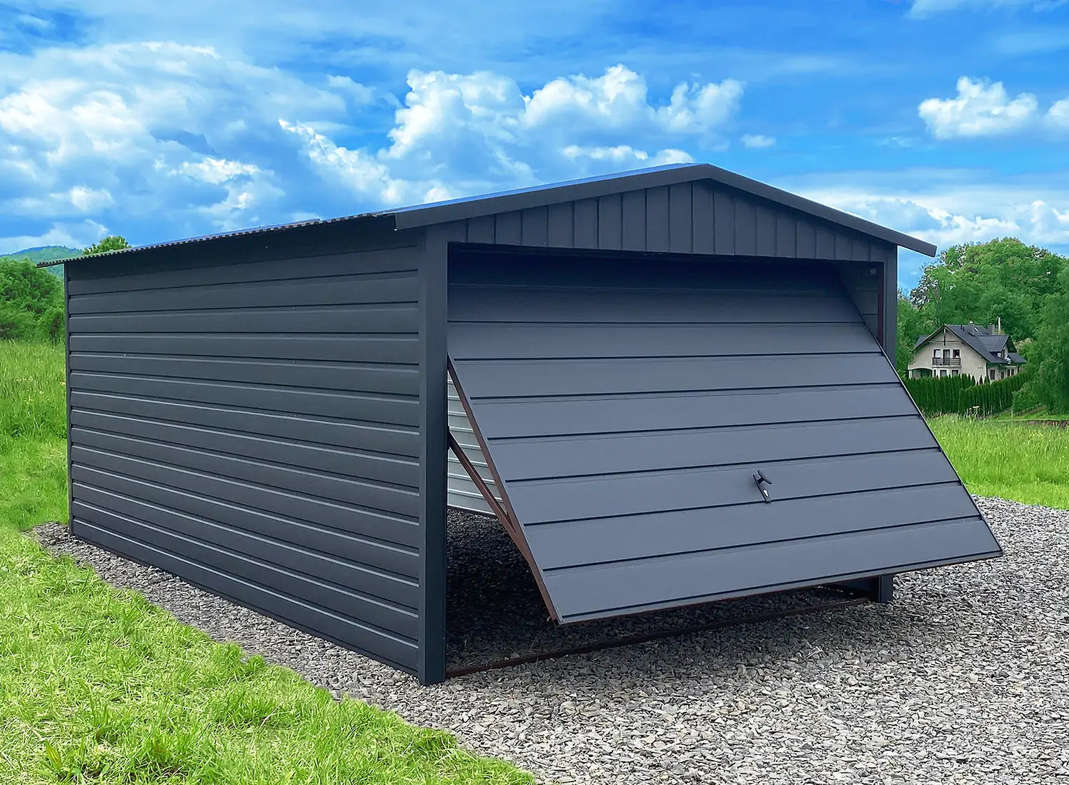 Grafitowy garaż blaszany z uchyloną bramą, na żwirowym podłożu, w tle zielony trawnik i dom. Błękitne niebo z chmurami w tle.