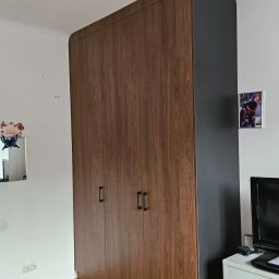 Łukasz Kołodziński - Wysoka, brązowo-czarna szafa trzydrzwiowa z zaokrąglonymi krawędziami, na drewnianej podłodze, z fragmentem biurka i telewizora po prawej stronie.