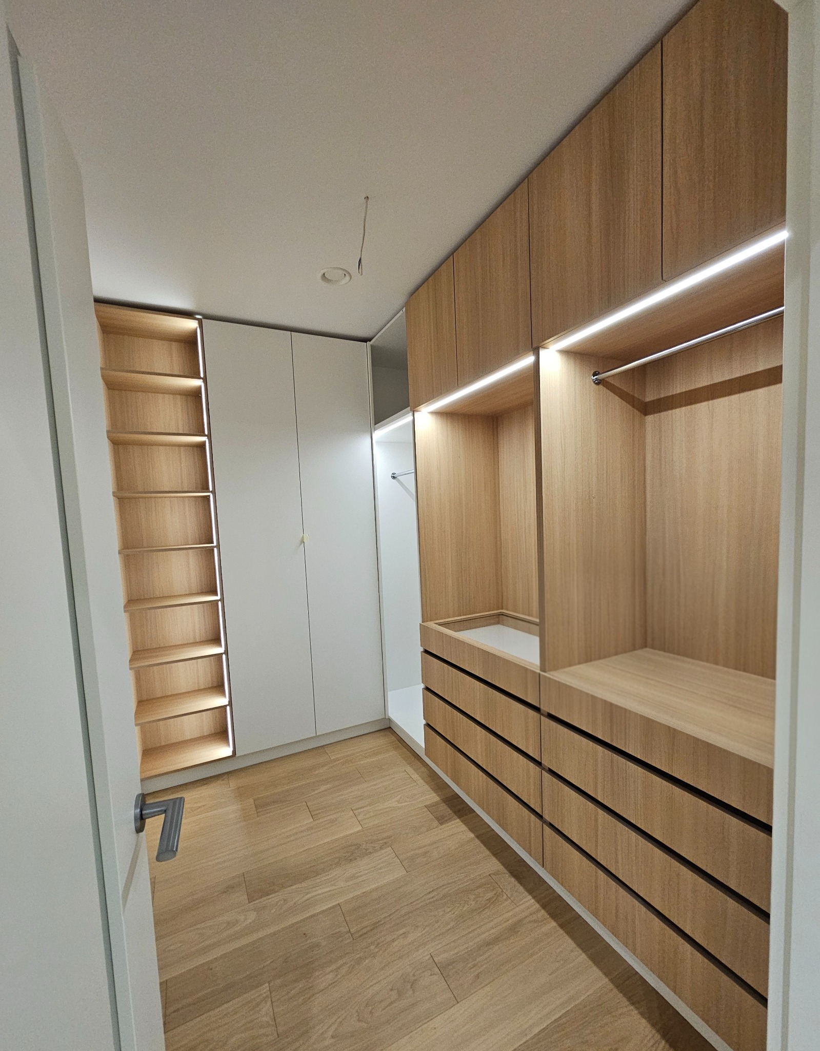 Jasna garderoba z oświetleniem LED: drewniane półki i szuflady po prawej, biała szafa z półkami po lewej. Parkiet na podłodze, widok z wnętrza pomieszczenia.