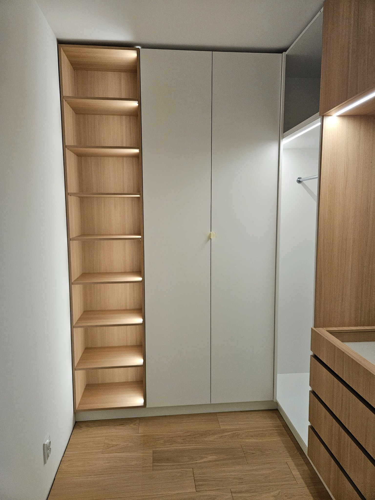 Nowoczesna szafa wnękowa z oświetleniem LED, jasne drewno i białe fronty, minimalistyczny design, widok z perspektywy podłogi, ciepłe światło podkreśla strukturę drewna.