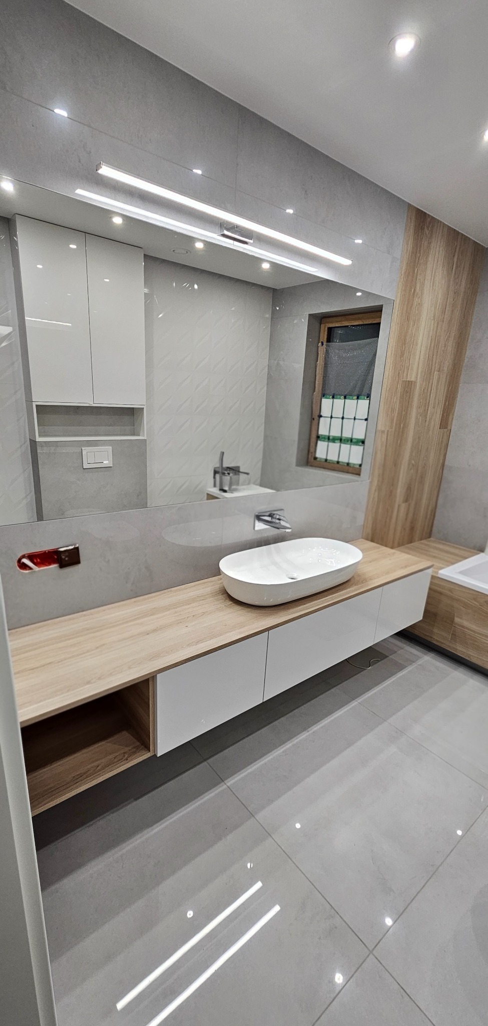Nowoczesna łazienka z owalną umywalką nablatową, szafką z drewnianym blatem i białymi frontami. W tle lustro z oświetleniem LED i szare płytki z subtelnym wzorem.
