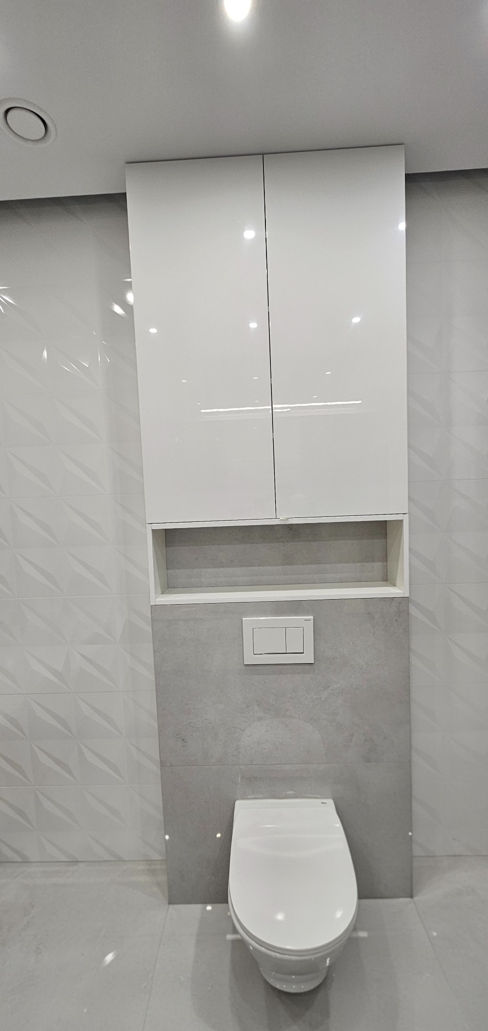 Nowoczesna toaleta podwieszana z szafką wnękową w minimalistycznej łazience. Białe fronty szafek i szara ściana tworzą spójną całość. Widoczny przycisk spłukiwania.