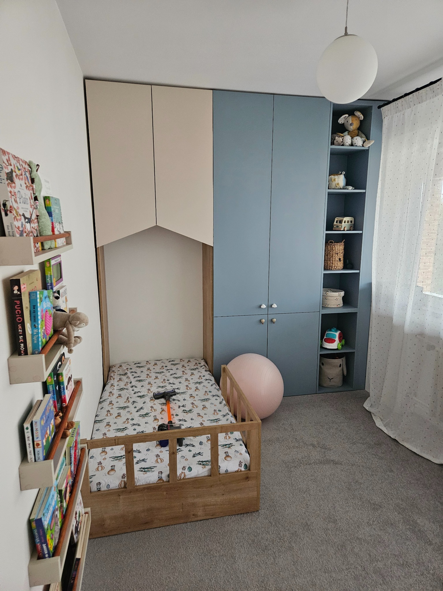 Pokój dziecięcy z zabudową meblową: szafa z półkami w odcieniach beżu i błękitu, łóżko z drewnianą ramą, półki na książki i dekoracje. Jasne wnętrze.