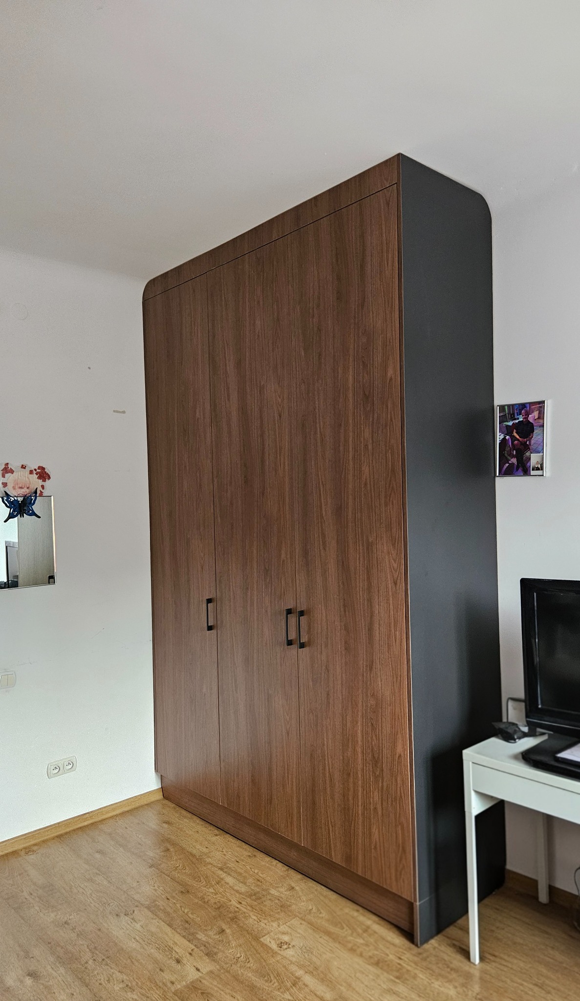 Wysoka, brązowo-czarna szafa trzydrzwiowa z zaokrąglonymi krawędziami, na drewnianej podłodze, z fragmentem biurka i telewizora po prawej stronie.