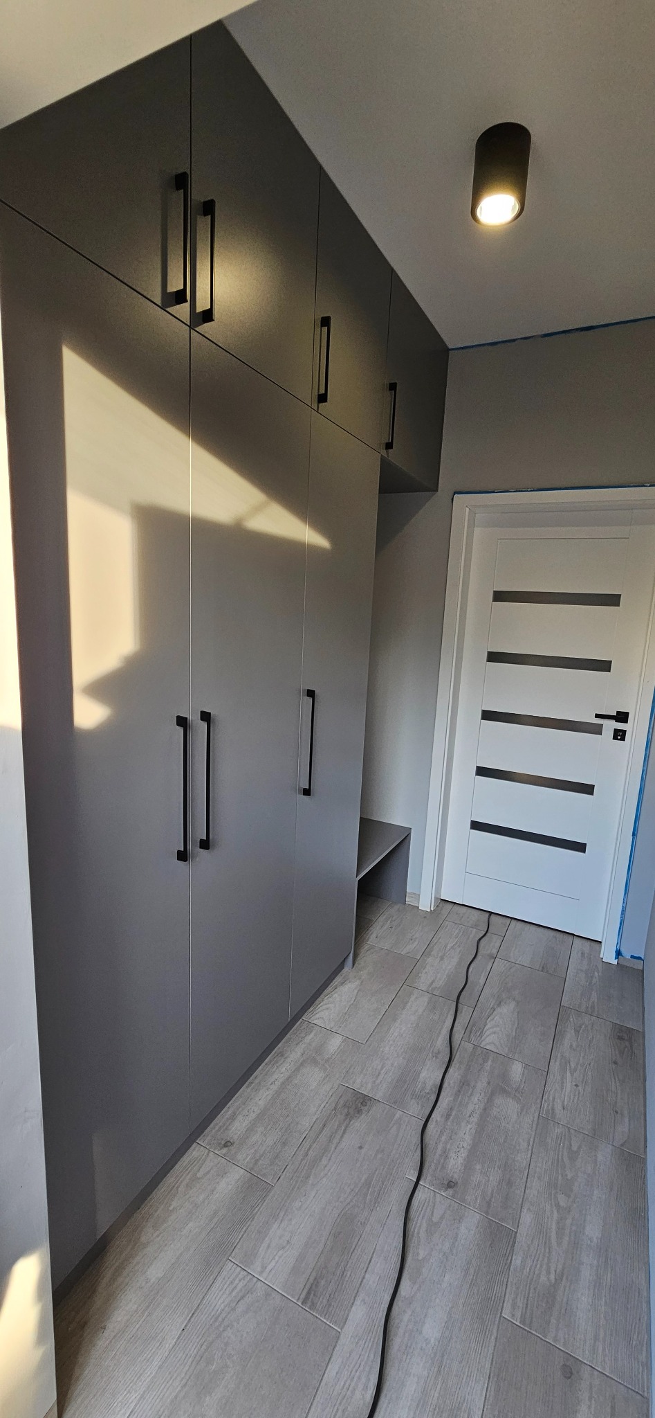 Szara zabudowa wnękowa z czarnymi uchwytami, dopasowana do wnętrza. Widoczna podłoga z paneli i białe drzwi z poziomymi przeszkleniami. Minimalistyczny design.