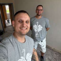 Pro Bau - Uśmiechnięty mężczyzna z tatuażami robi selfie na tle ściany po malowaniu. W tle drugi mężczyzna i otwarte drzwi. Widoczny fragment podłogi przed wykończeniem.