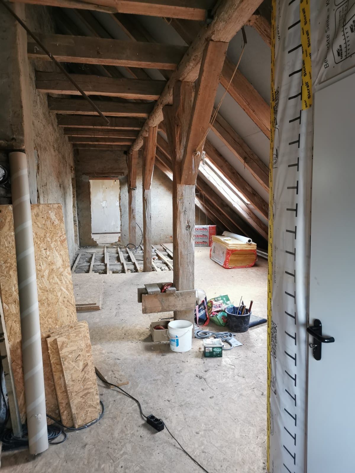 Poddasze w trakcie remontu: widoczne drewniane belki stropowe, częściowo ułożona podłoga, materiały izolacyjne i narzędzia. Prace wykończeniowe w surowym stanie.