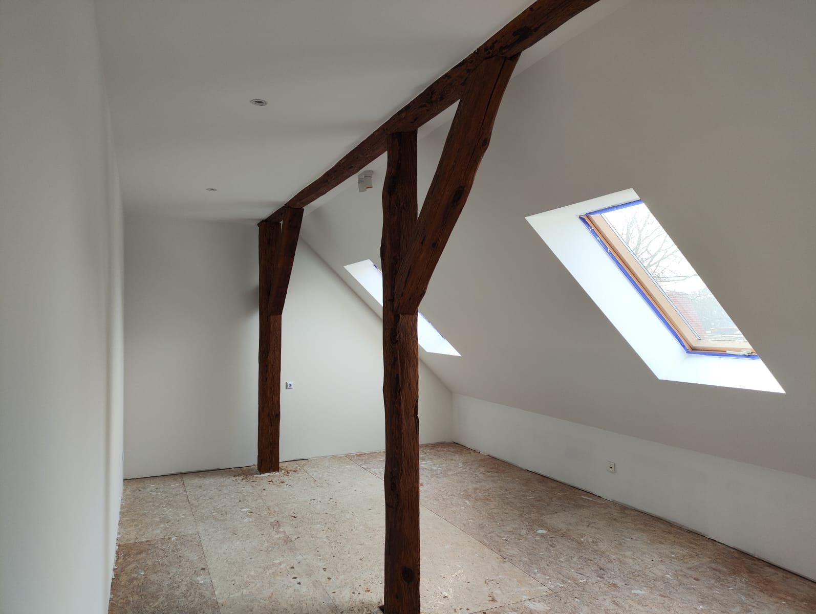 Białe poddasze z drewnianą konstrukcją nośną i oknami dachowymi. Świeżo pomalowane ściany, przygotowane do dalszych prac wykończeniowych.