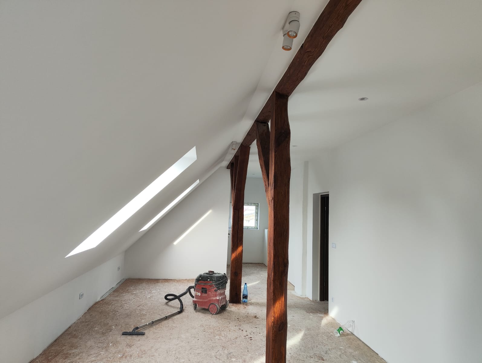 Białe poddasze po remoncie z drewnianymi belkami, oknami dachowymi i odkurzaczem przemysłowym na podłodze. Widoczne ślady po remoncie.
