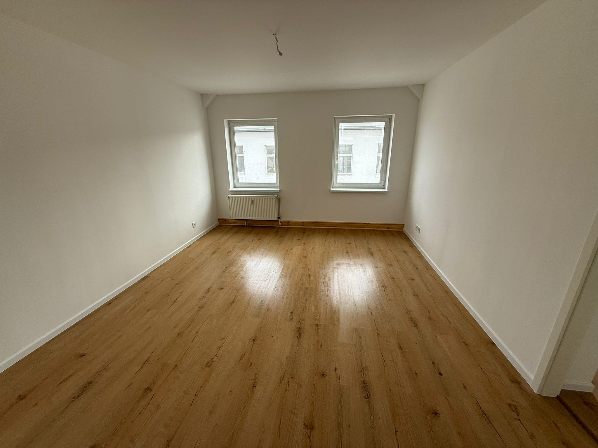 Pusty pokój po remoncie: świeżo pomalowane białe ściany, panele podłogowe imitujące drewno, dwa okna z grzejnikiem. Widoczne gniazdka elektryczne i wystający kabel z sufitu.