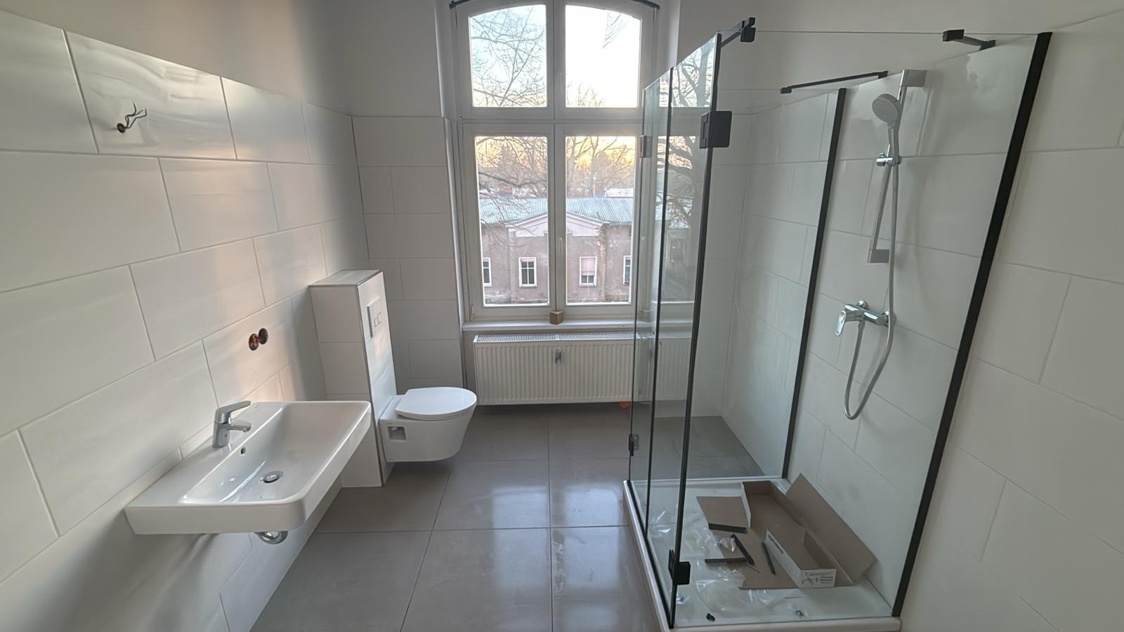 Jasna łazienka po remoncie: białe płytki, kabina prysznicowa ze szkła, umywalka i toaleta w nowoczesnym stylu. Widok na budynek przez okno.