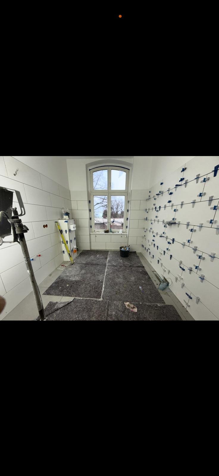 Biała łazienka w trakcie remontu. Na ścianach ułożone białe płytki z niebieskimi krzyżykami dystansowymi. Podłoga zabezpieczona szarą włókniną.