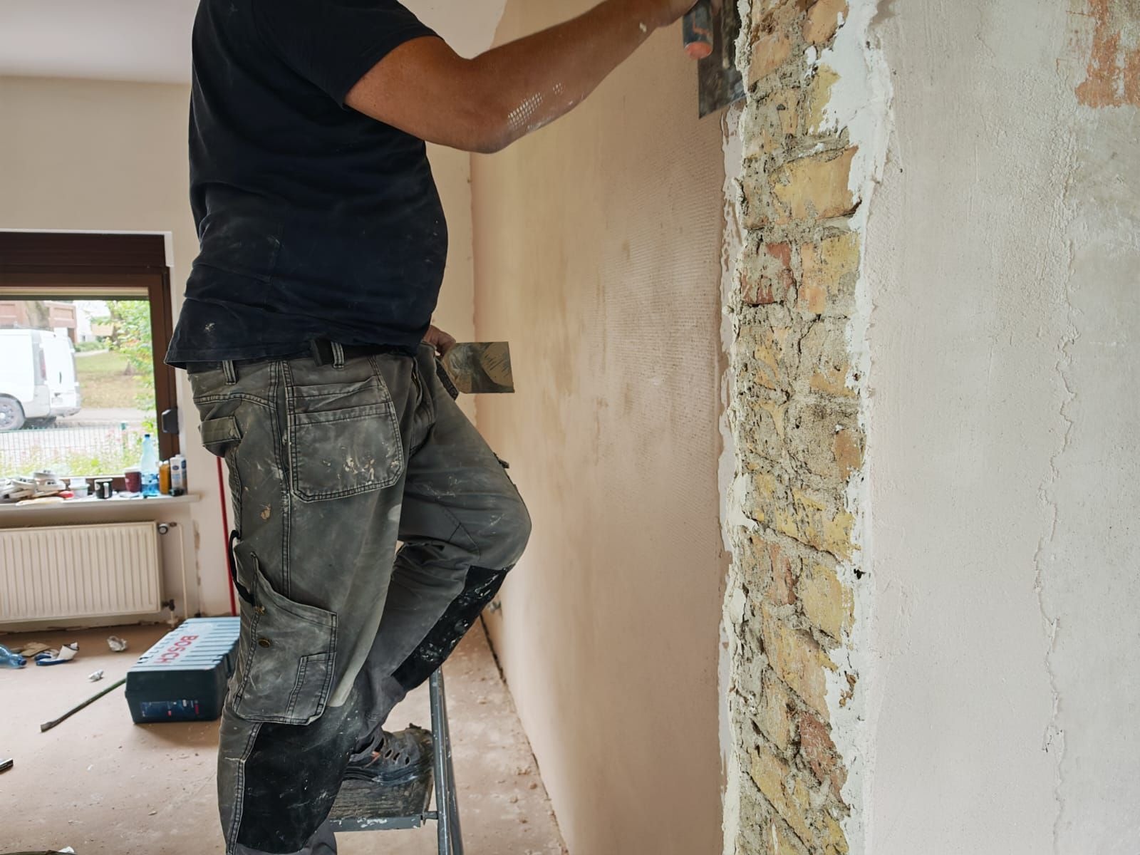 Mężczyzna na drabinie szpachluje ścianę z fragmentem odsłoniętej cegły. Widoczna skrzynka narzędziowa Bosch i okno w tle. Prace wykończeniowe w trakcie.