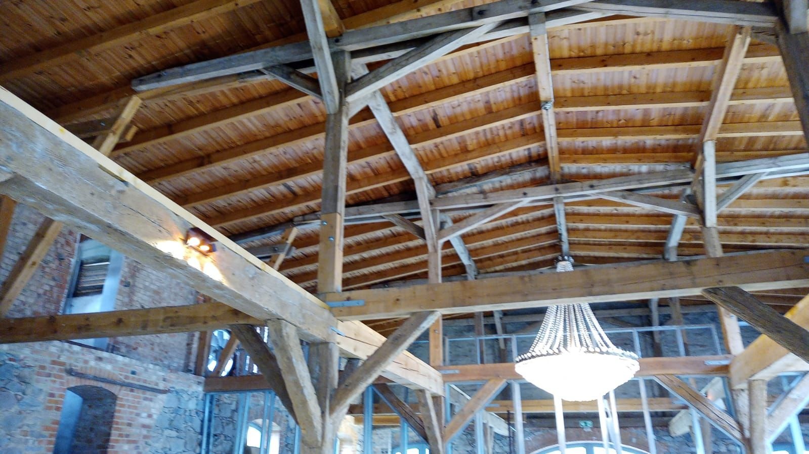 Drewniana konstrukcja dachu z widocznymi belkami i deskami, oświetlona lampą. W tle fragment murowanej ściany i okna. Widok z dołu.