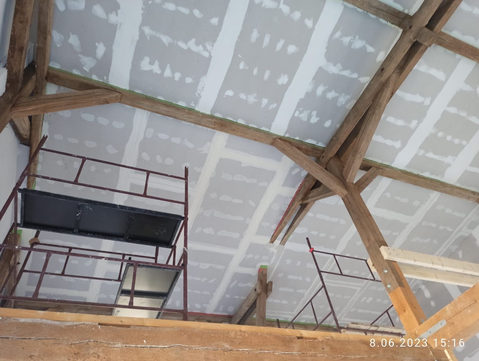 Montaż płyt karton-gips na drewnianej konstrukcji dachu, widok z dołu. Widoczne rusztowania i częściowo zakryte łączenia płyt. Praca w toku.