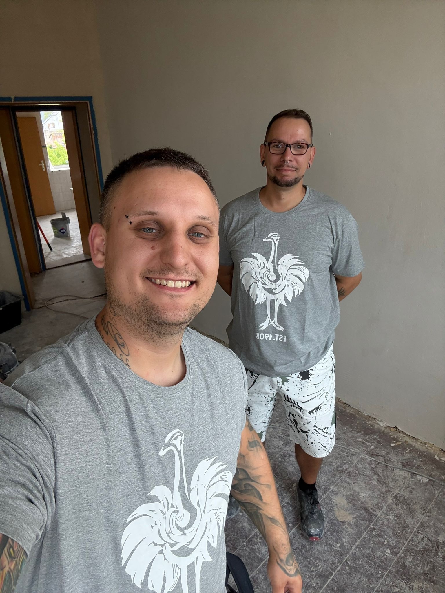 Uśmiechnięty mężczyzna z tatuażami robi selfie na tle ściany po malowaniu. W tle drugi mężczyzna i otwarte drzwi. Widoczny fragment podłogi przed wykończeniem.