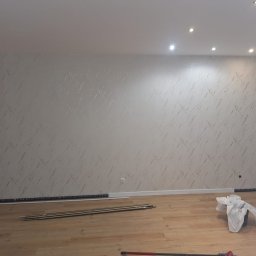 Nowy horyzont - Tapeta w jasnym pokoju z drewnianą podłogą. Widoczne listwy przypodłogowe i narzędzia. Ściana pokryta tapetą o delikatnym wzorze w odcieniach bieli i złota.