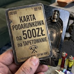 Nowy horyzont - Karta podarunkowa na 500 zł na tapetowanie, trzymana w dłoni. W tle zapalniczki i fragment sofy. Karta w stylu górniczym z motywem kopalni.
