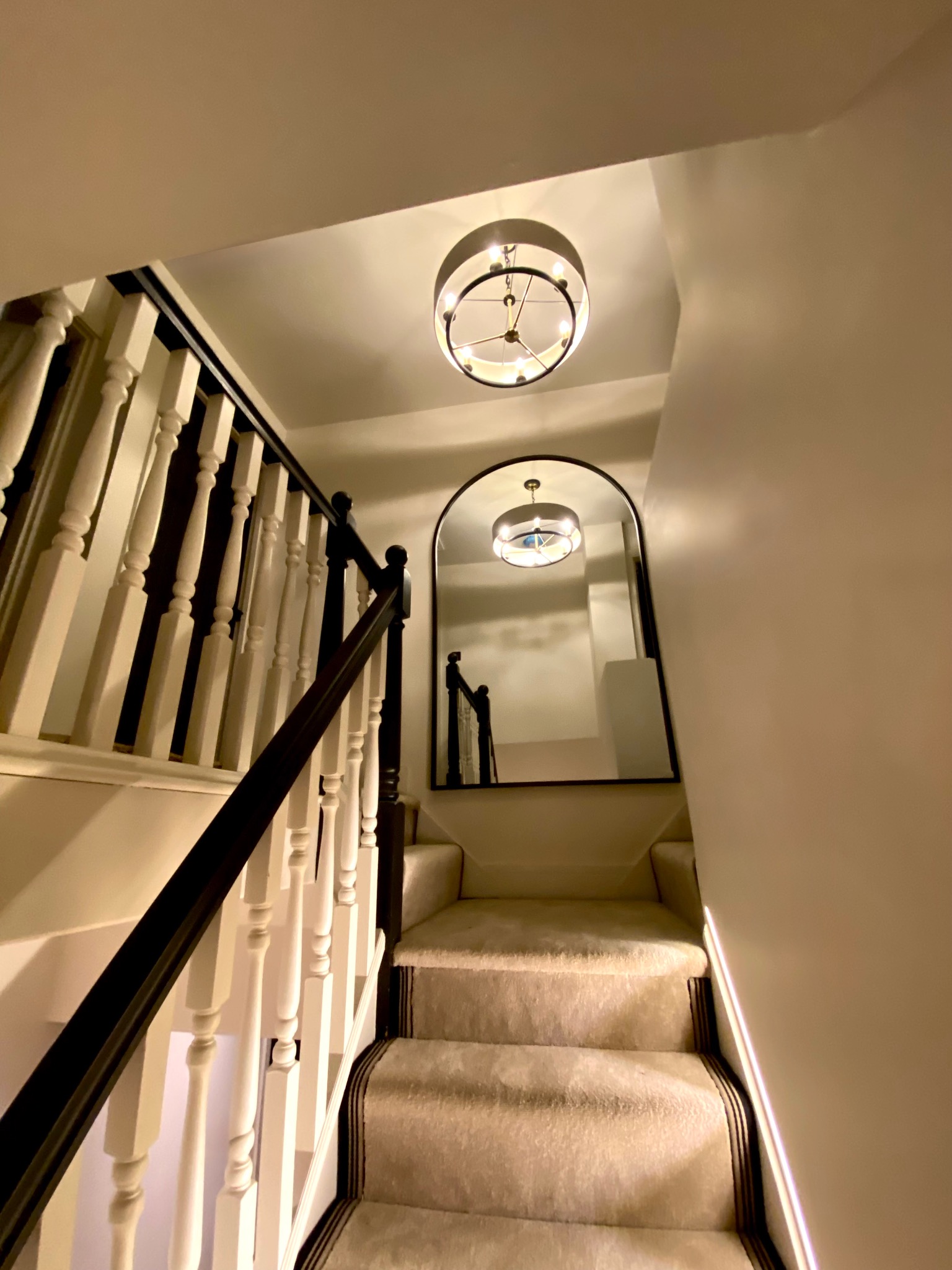 Klatka schodowa z beżową wykładziną, białą balustradą z czarnym poręczą i okrągłą lampą. W lustrze odbija się lampa i fragment schodów.