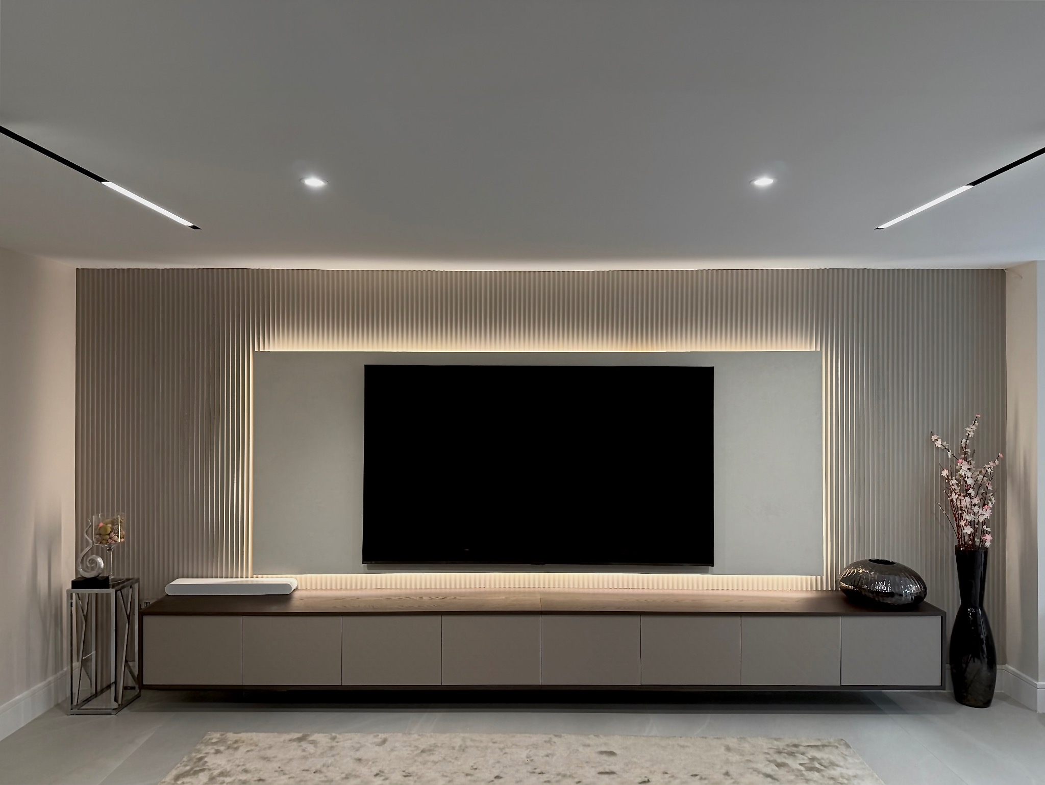 Nowoczesna ściana TV z podświetlanymi lamelami i szafką RTV w odcieniach beżu. Minimalistyczny design z dekoracjami i dywanem w tle.