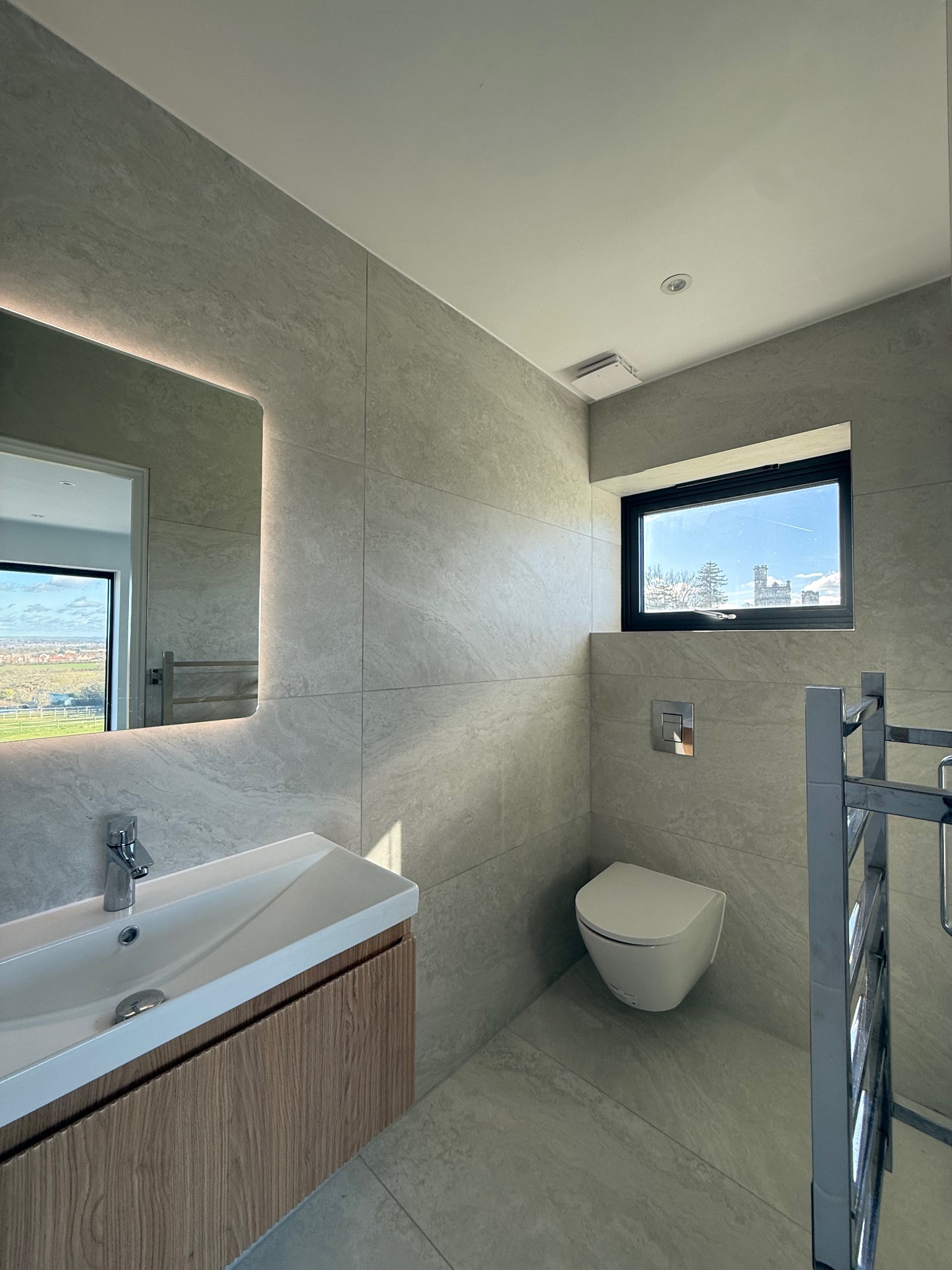 Nowoczesna łazienka z szarymi płytkami, podświetlanym lustrem, wiszącą toaletą i drewnianą szafką pod umywalką. Okno z widokiem na krajobraz.