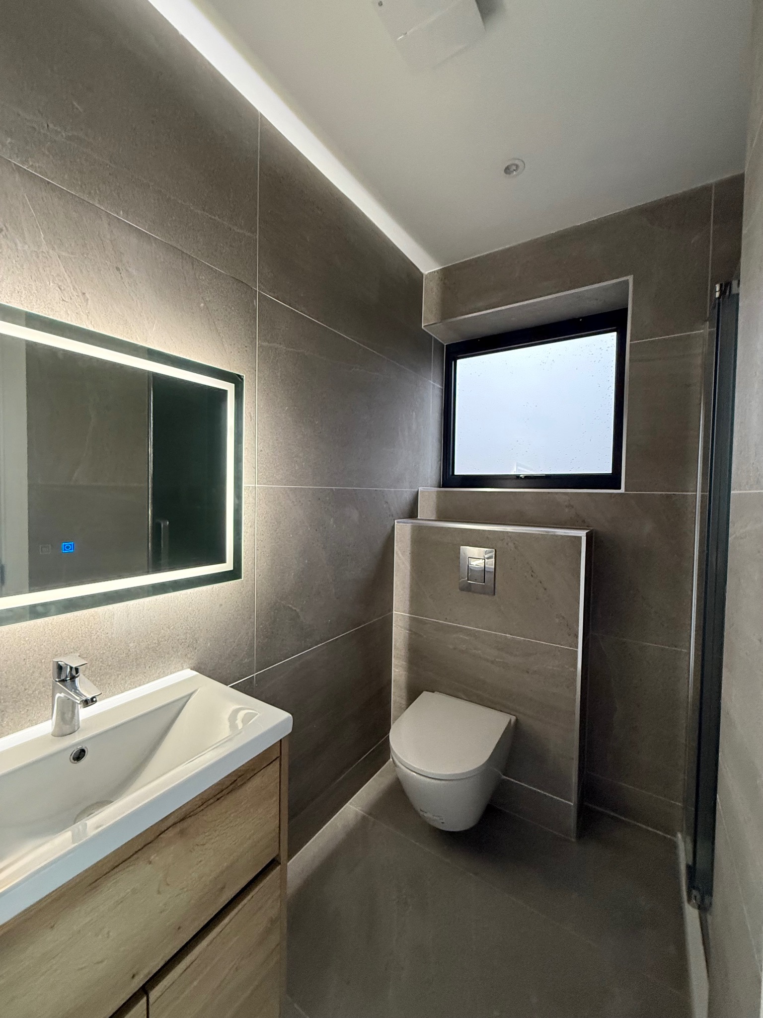 Nowoczesna łazienka z szarymi płytkami, podświetlanym lustrem nad umywalką z drewnianą szafką i wiszącą toaletą. Okno z czarną ramą doświetla wnętrze.