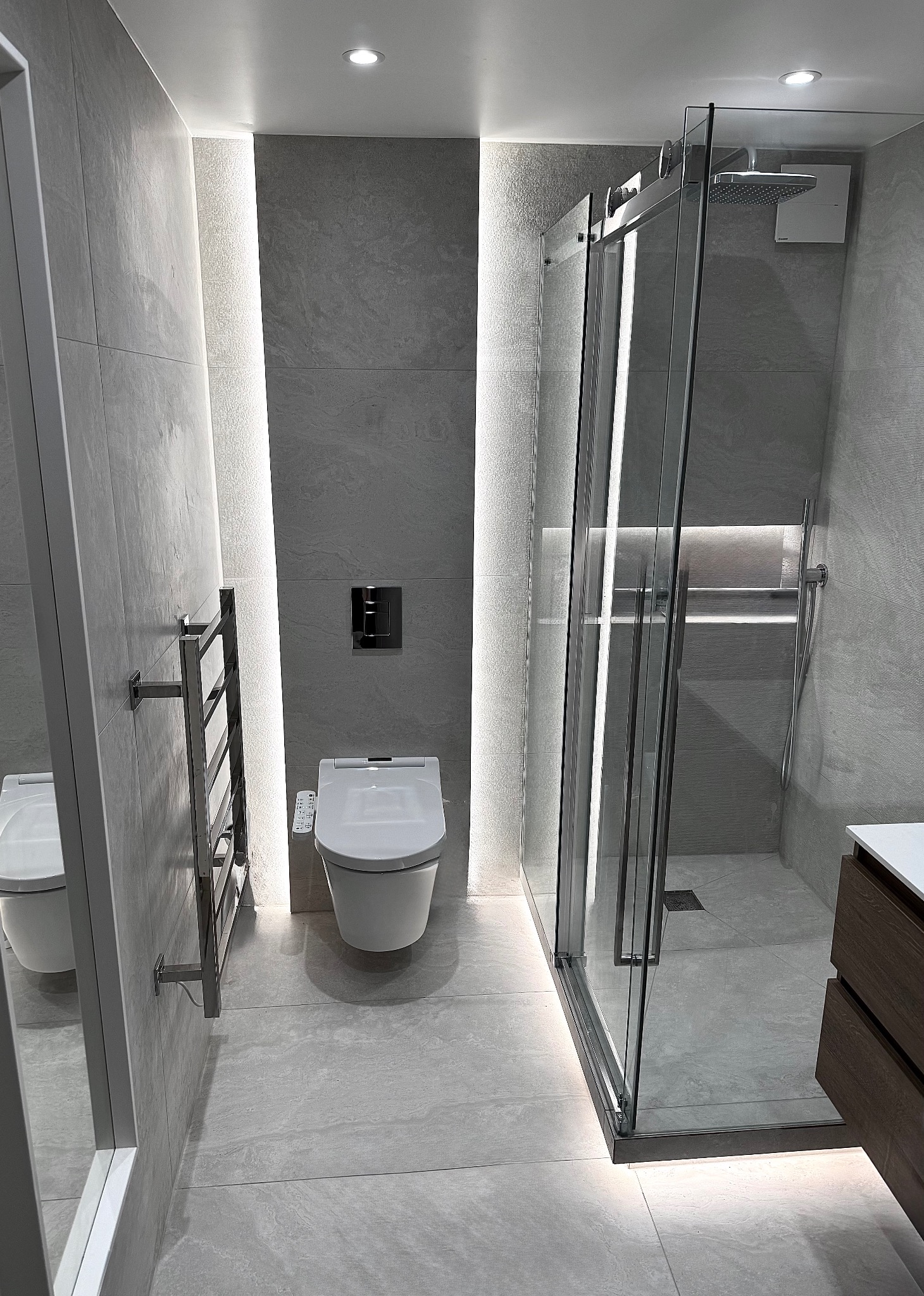 Nowoczesna łazienka w szarościach z kabiną prysznicową typu walk-in, podświetleniem LED i wiszącą toaletą z panelem sterowania. Minimalistyczny design z chromowanymi akcentami.