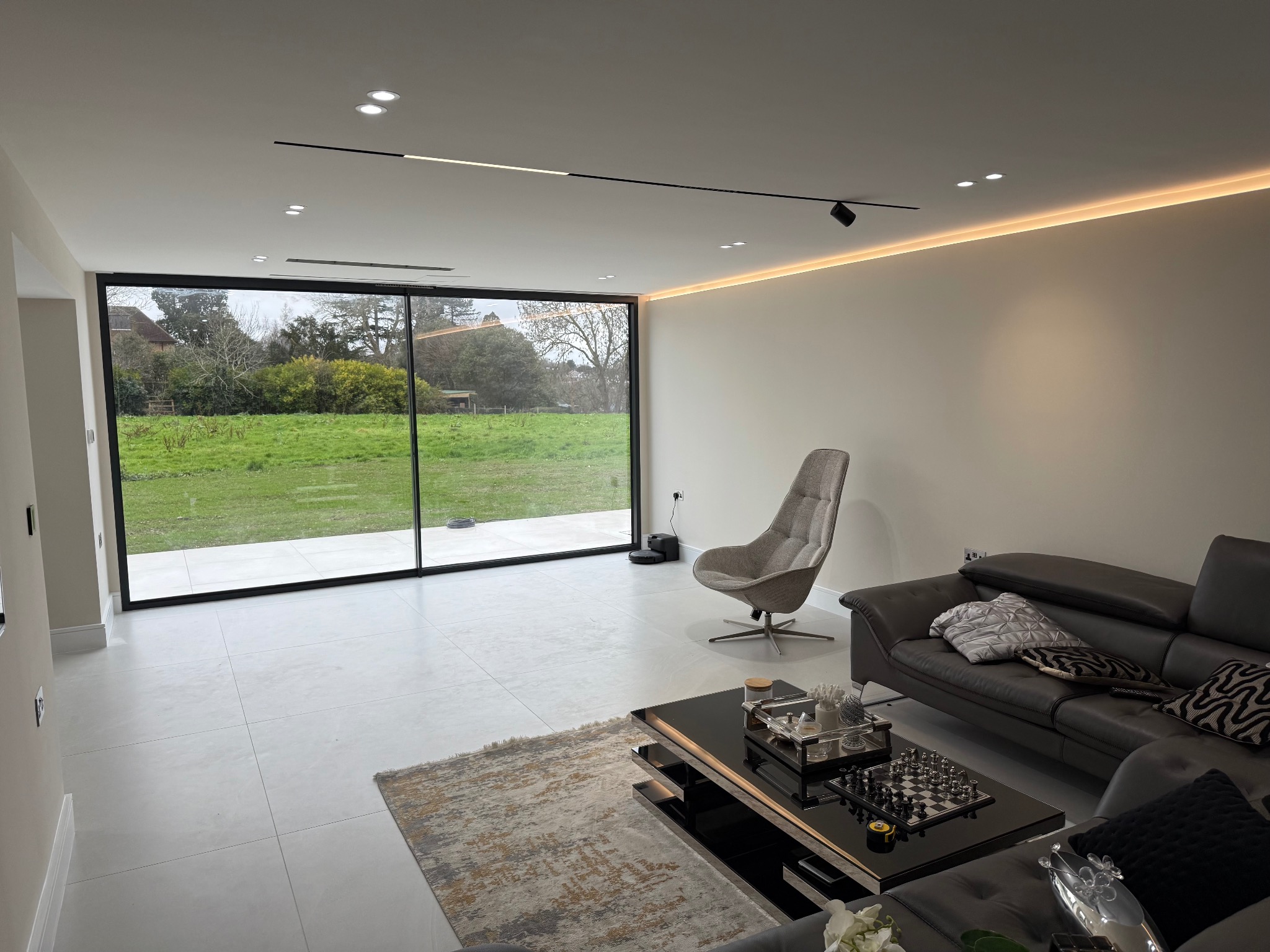 Nowoczesny salon z dużym oknem na ogród. Minimalistyczny design, szara sofa, fotel i dywan. Podwieszany sufit z oświetleniem LED. Szachownica na stoliku kawowym.