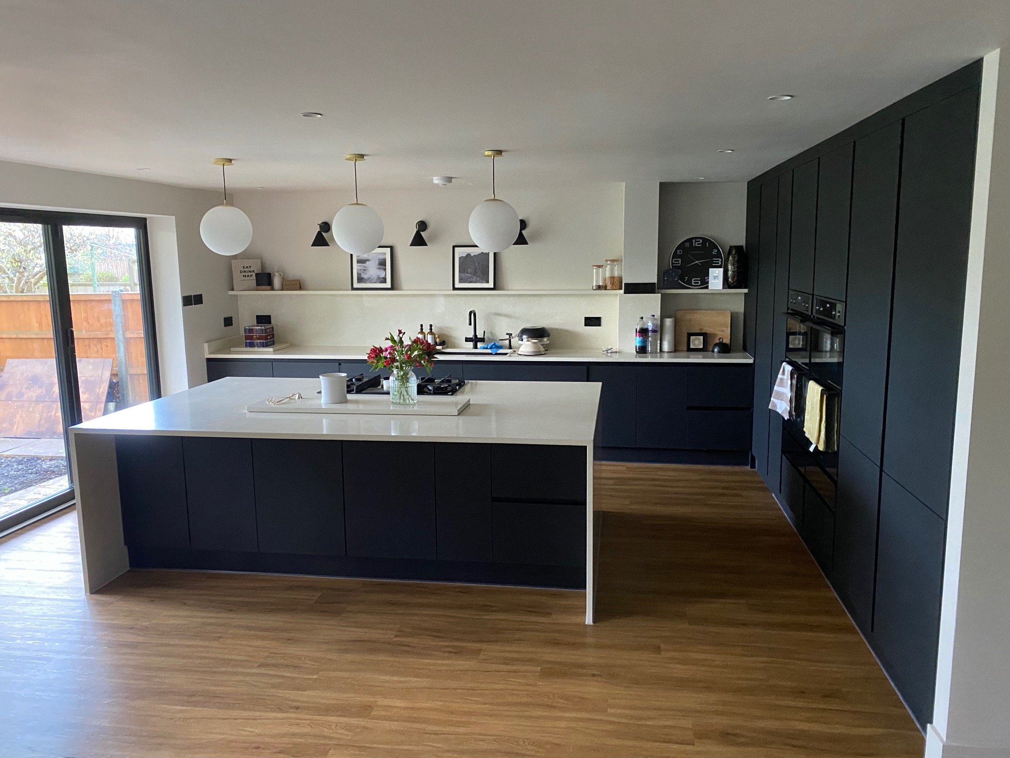 Nowoczesna kuchnia z wyspą i zabudową w kolorze grafitu. Jasny blat, drewniana podłoga, okrągłe lampy. Minimalistyczny design z akcentami w postaci kwiatów i dekoracji.