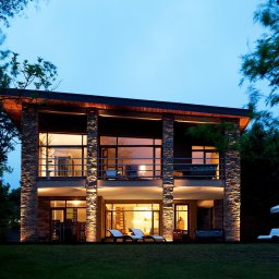Fabian Wacławski Inwestor Zastępczy - Dwukondygnacyjny dom z kamiennymi kolumnami oświetlonymi od dołu, z dużymi oknami i balkonem, otoczony zielenią, sfotografowany o zmierzchu.