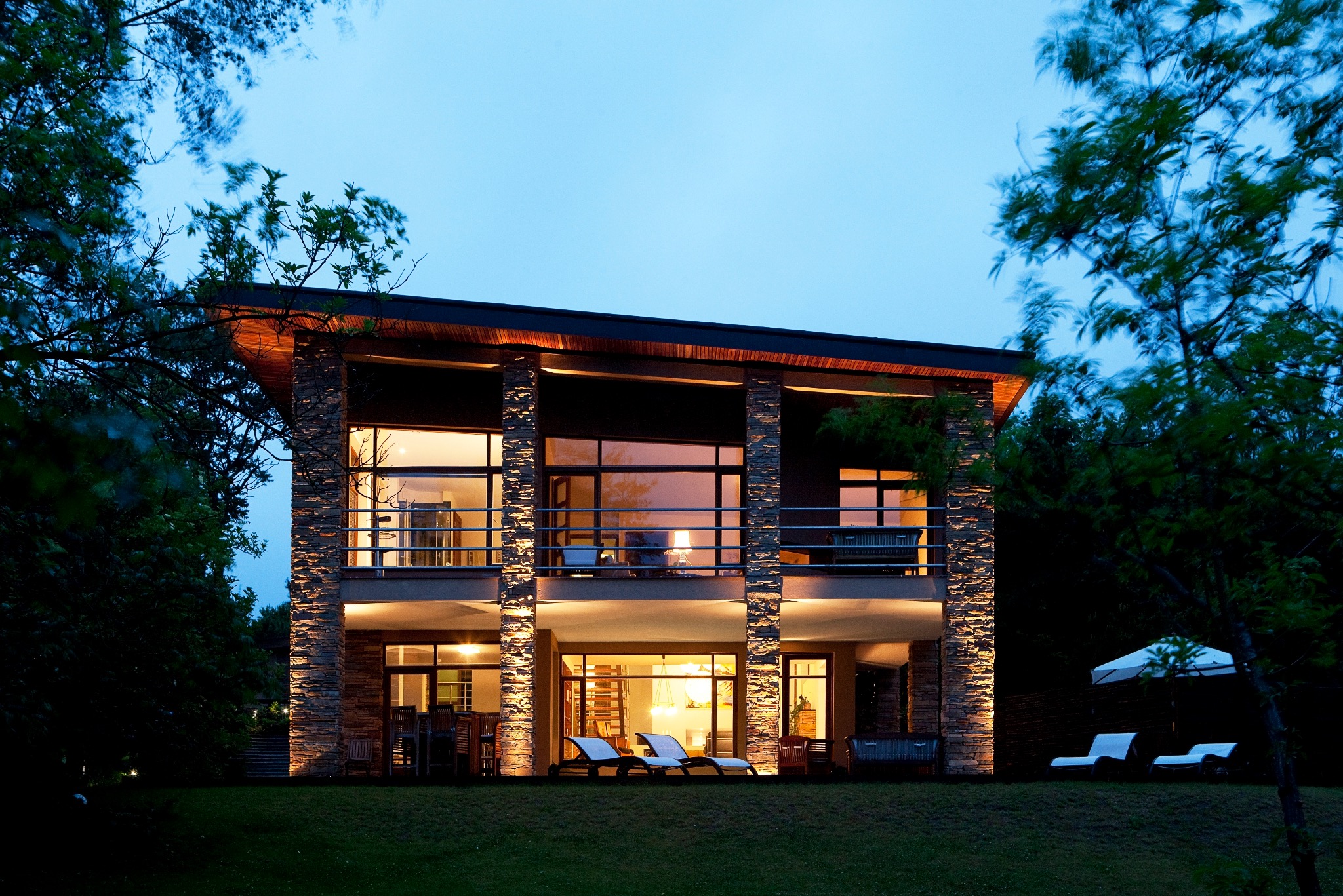 Dwukondygnacyjny dom z kamiennymi kolumnami oświetlonymi od dołu, z dużymi oknami i balkonem, otoczony zielenią, sfotografowany o zmierzchu.