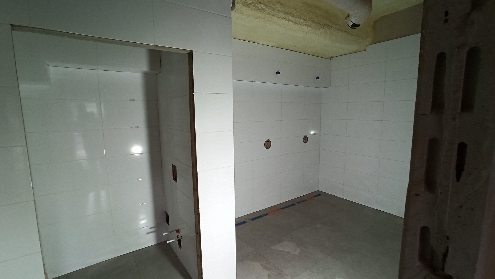 Białe płytki ścienne w surowym wnętrzu łazienki, widoczne otwory na instalacje i fragment sufitu z pianką montażową. Częściowo wykończone pomieszczenie.
