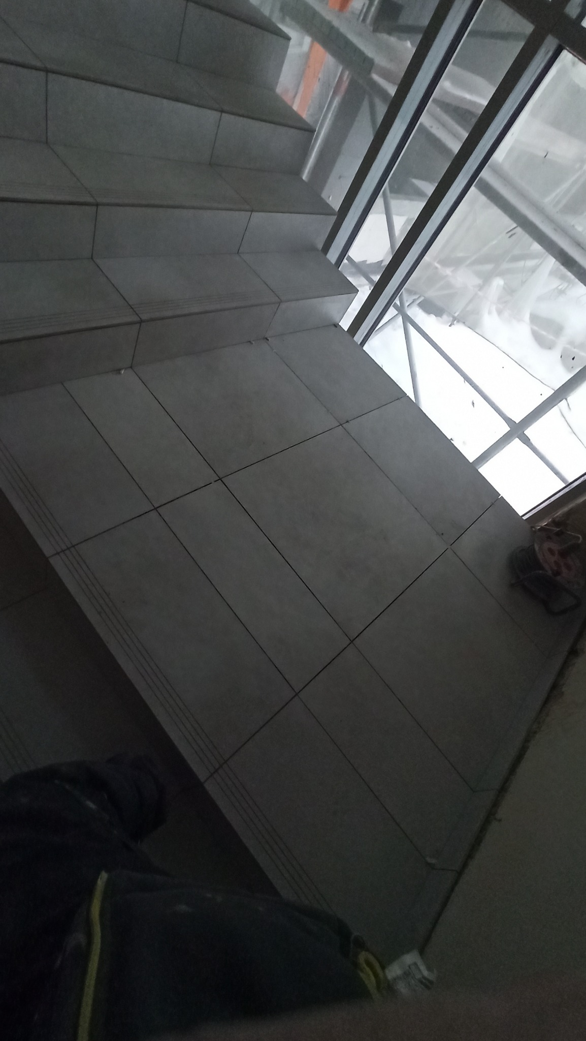 Wykończone płytkami schody wewnątrz budynku, widoczne przez okno elementy konstrukcji. Płytki w odcieniach szarości, ułożone równo. Ujęcie z dołu, z fragmentem ubrania w rogu.