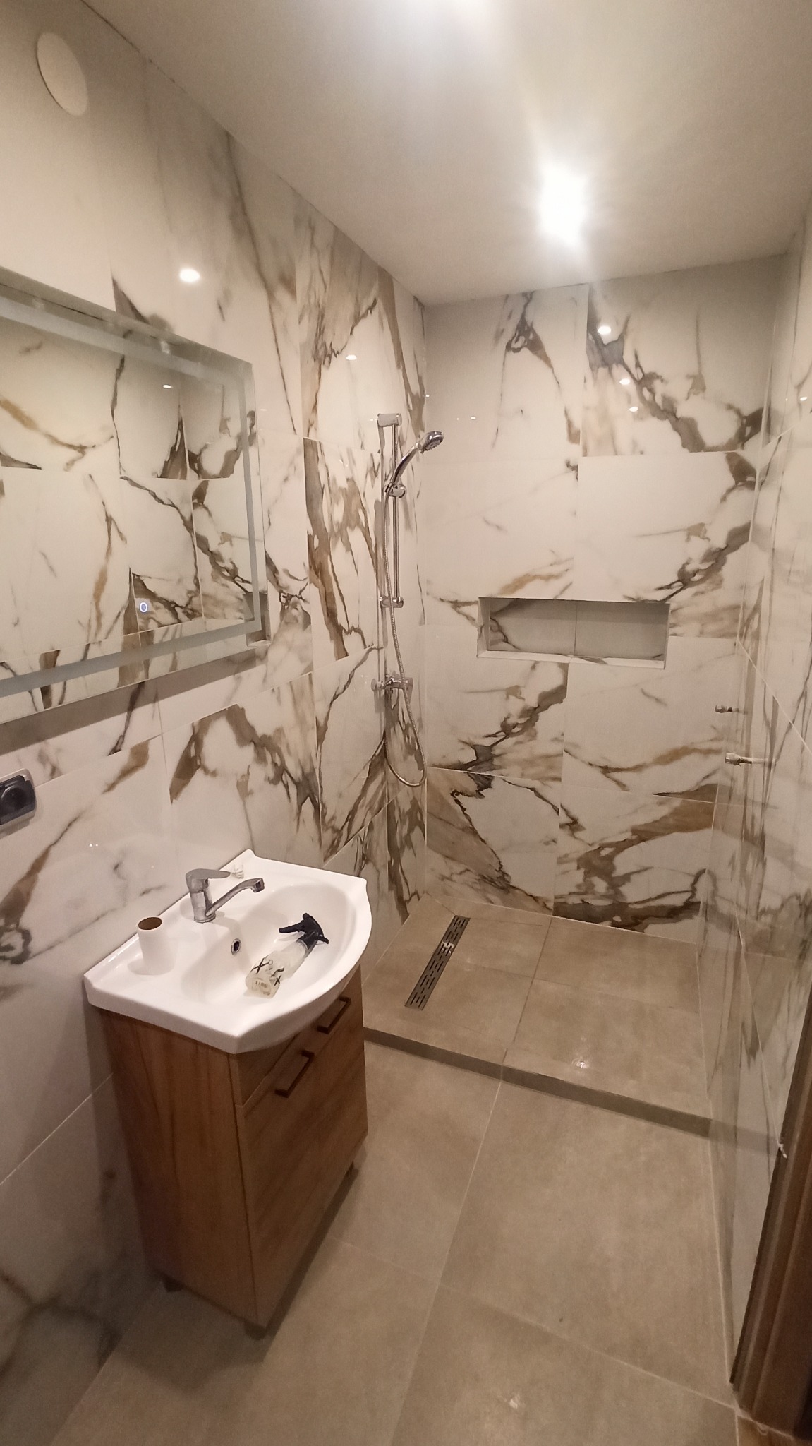 Wykończona łazienka z kabiną prysznicową typu walk-in, umywalką z szafką z drewna i lustrem. Ściany i podłoga wyłożone płytkami imitującymi marmur w odcieniach bieli i brązu.