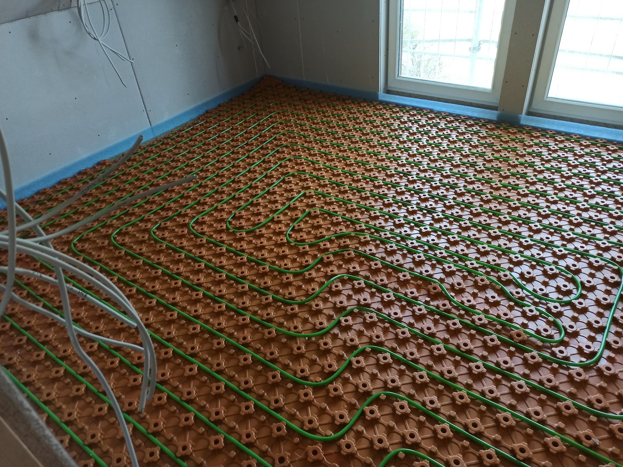 Ułożona instalacja ogrzewania podłogowego z zielonymi rurami na brązowej macie montażowej, widok z góry, kable elektryczne w rogu, przygotowanie do wylewki.