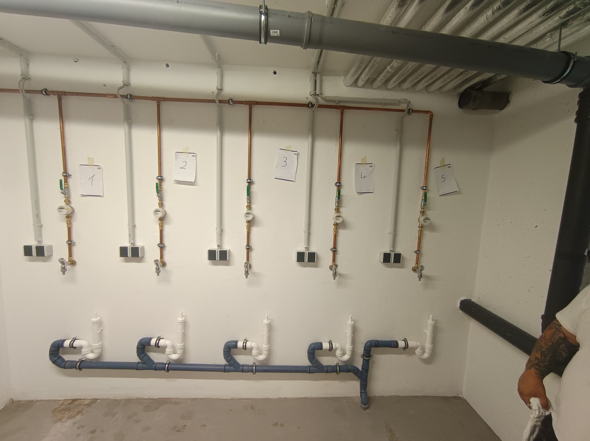 Rzędy rur miedzianych i PCV z licznikami, ponumerowane od 1 do 5, zamontowane na białej ścianie w piwnicy. Widoczne elementy instalacji wodnej i kanalizacyjnej.