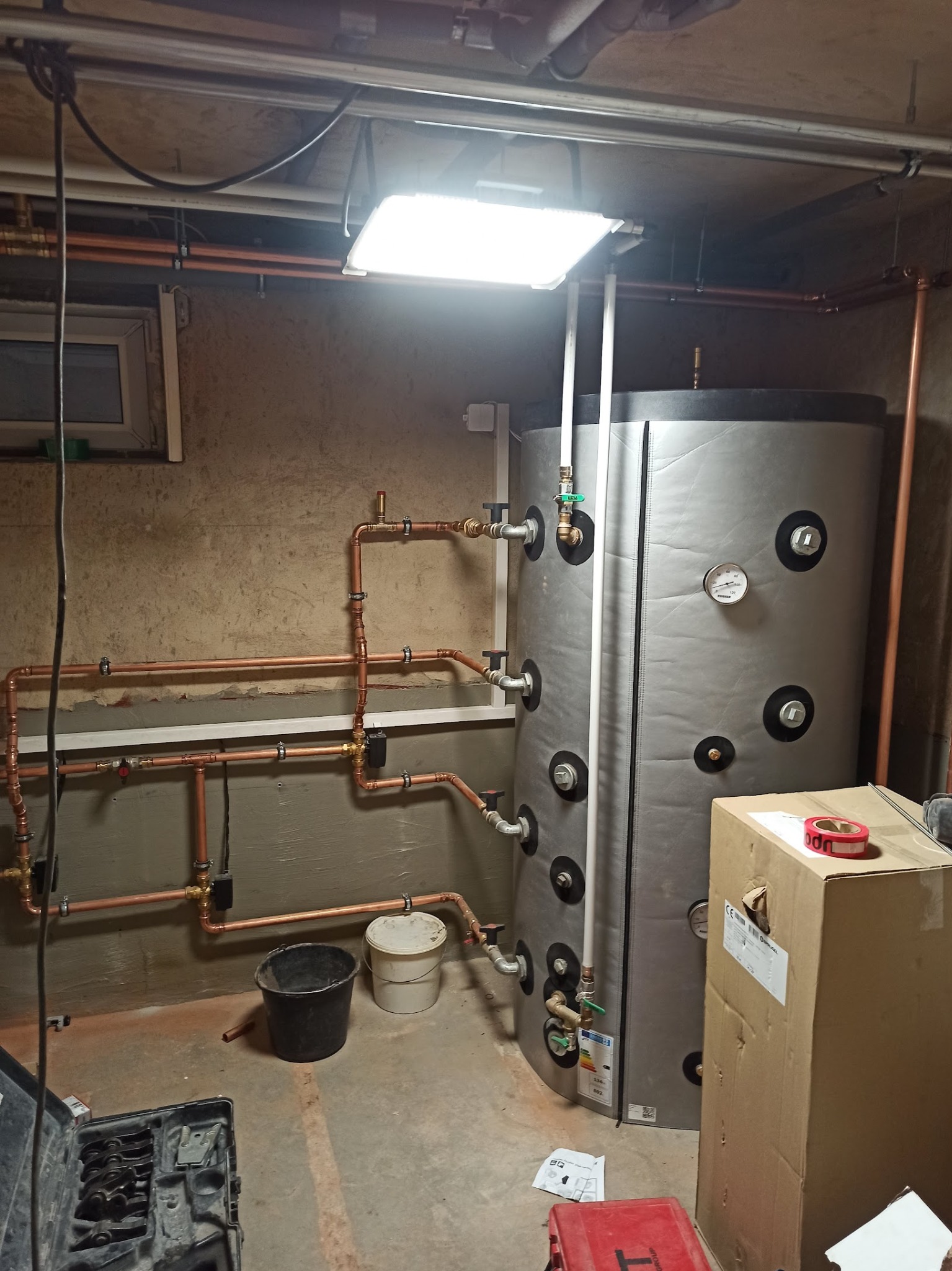 Instalacja bojlera z miedzianą armaturą w piwnicy w Świdnicy. Widoczne rury, zbiornik i narzędzia hydrauliczne. Oświetlenie sufitowe.