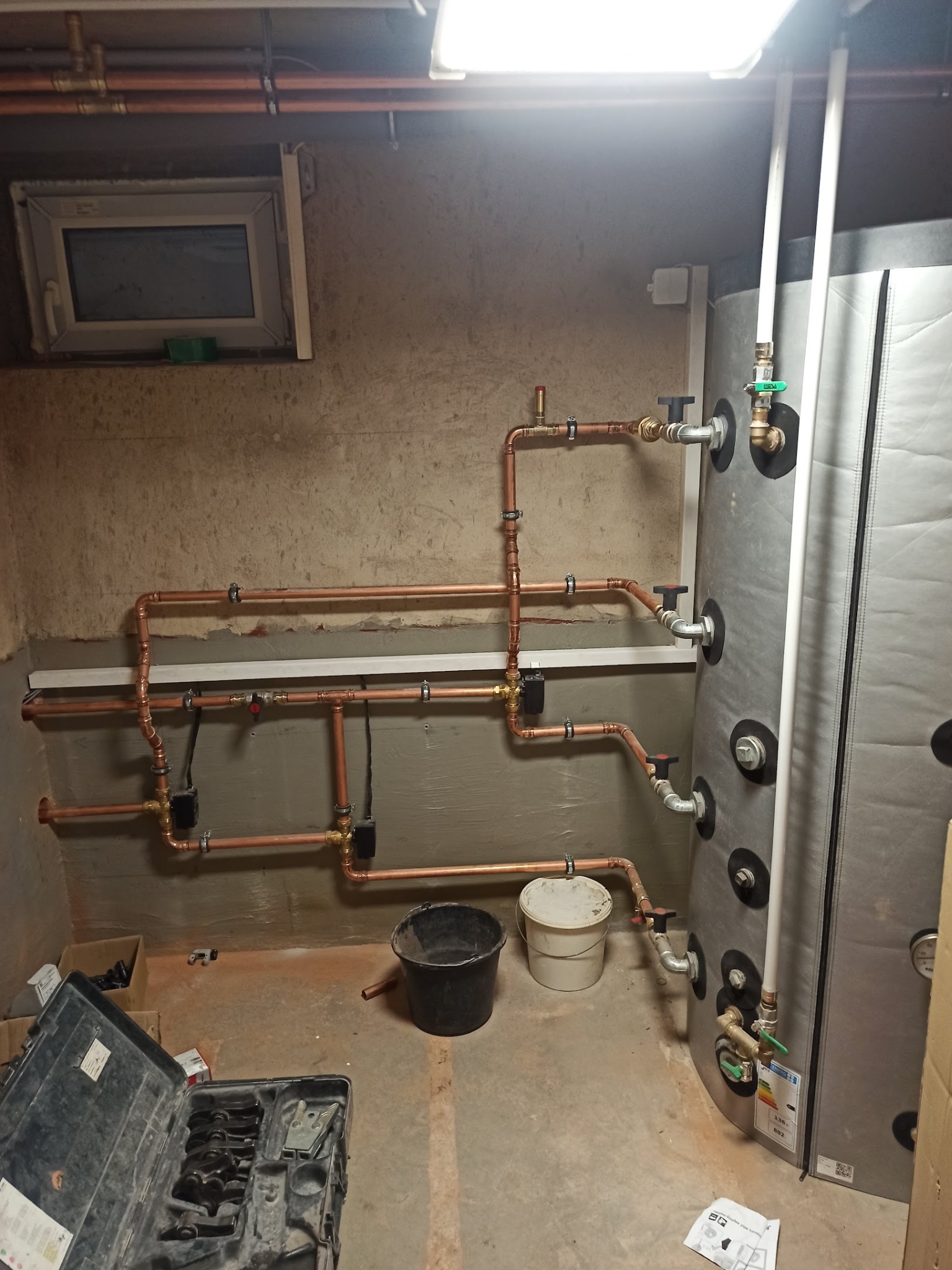 Miedziana instalacja grzewcza z zbiornikiem akumulacyjnym w piwnicy, widoczne rury, zawory i narzędzia hydrauliczne. Surowe ściany i podłoga.
